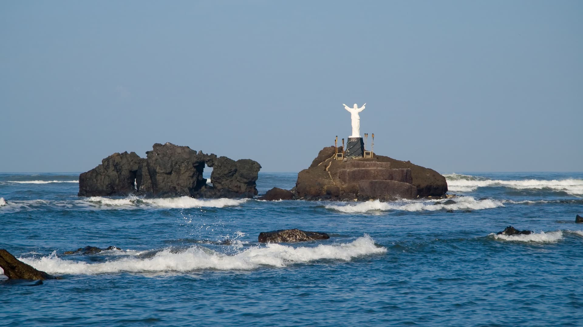 A statue of Jesus stands on a rocky islet in the Pacific Ocean, with waves crashing around it near the port of Acajutla, El Salvador.