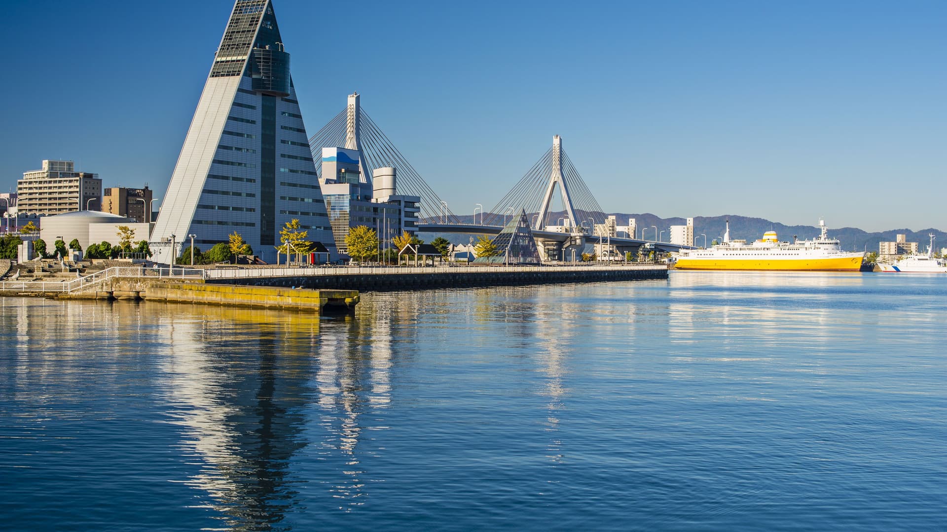 A wide, scenic view of the Aomori Bay Bridge and the iconic pyramid-shaped ASPAM building, with a large orange-and-white ferry docked in the harbor.
