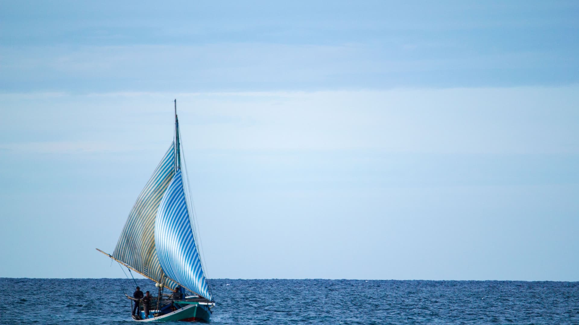 A sailboat with blue and white striped sails sailing on the calm blue waters of the Indian Ocean near Ashmore Reef, Australia, on a hazy day.