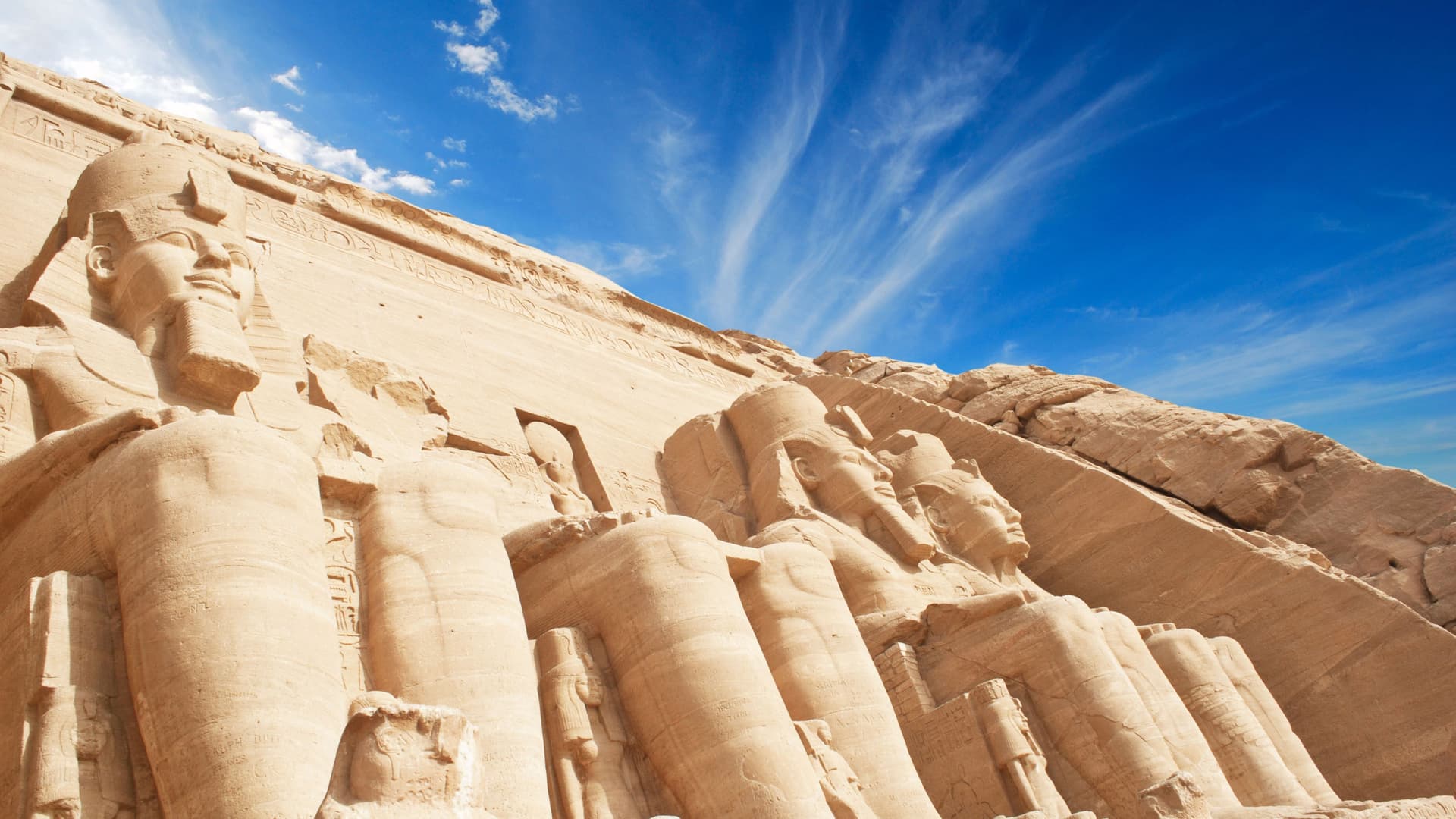 A stunning wide-angle view of the colossal rock-carved statues of Ramses II at the ancient Egyptian temple of Abu Simbel, near Aswan, Egypt, under a bright blue sky.
