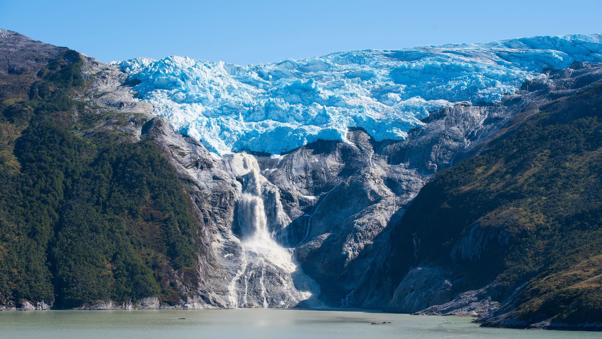 A large glacier on a mountain cliff, with a powerful waterfall cascading down into a fjord in the Chilean Fjords.