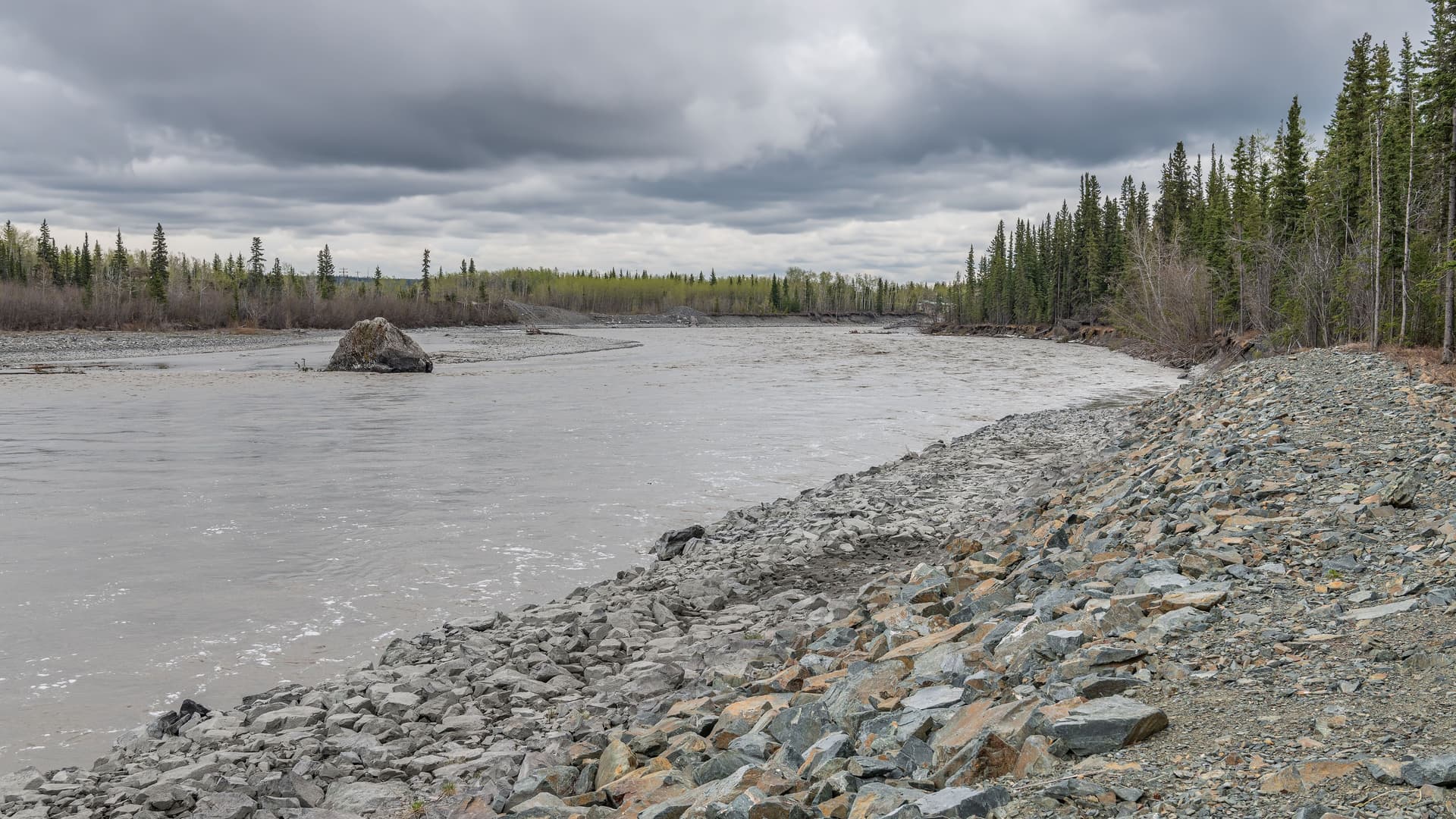 "A view of the wide, gray-colored Copper River in Alaska, with a rocky shore and evergreen trees lining the banks under a dramatic, cloudy sky. "