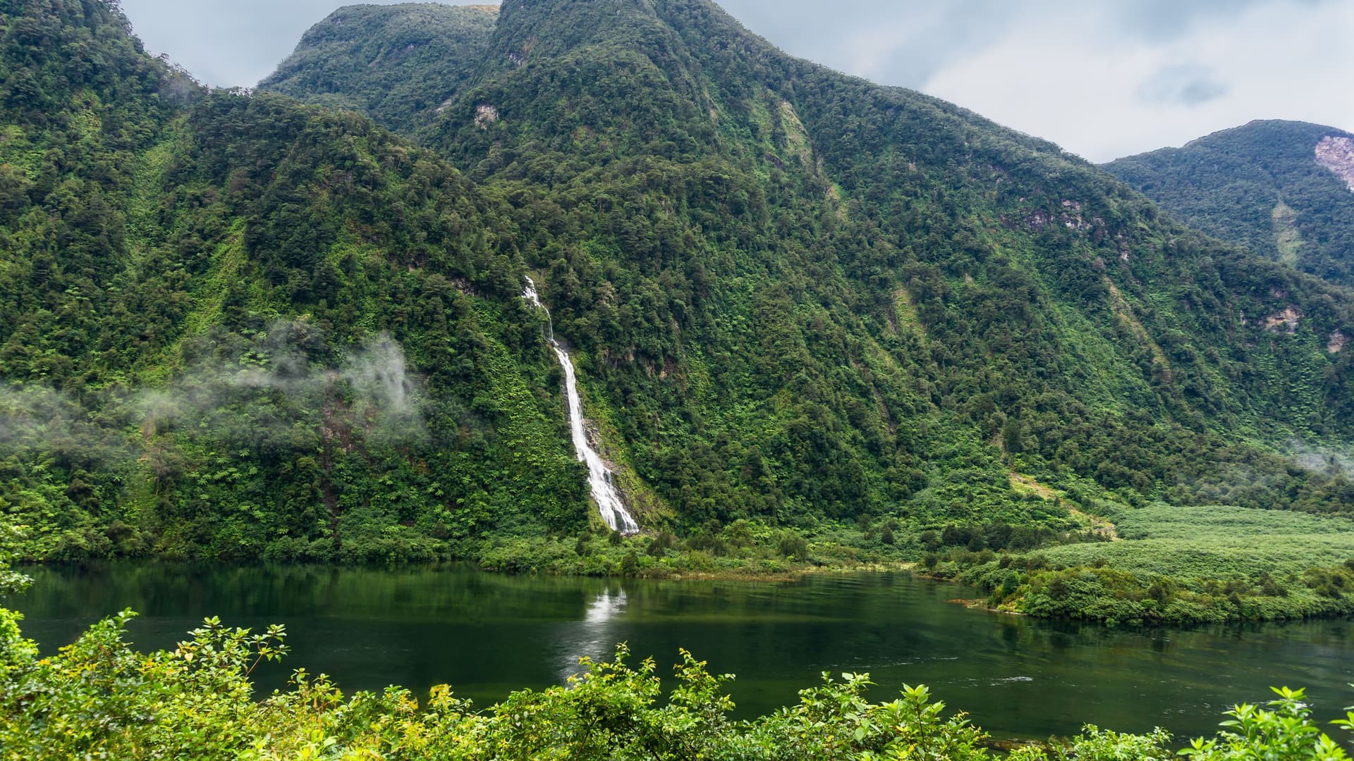 The stunning, mist-shrouded landscape of Doubtful Sound, New Zealand, with a powerful waterfall cascading down a dense, rainforest-covered mountain into the fjord.