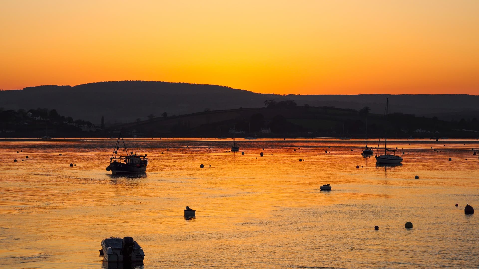 A tranquil sunset view over the Exe Estuary in Exmouth, Devon, with fishing boats and sailboats dotting the calm, reflective orange water and hills silhouetted in the background.