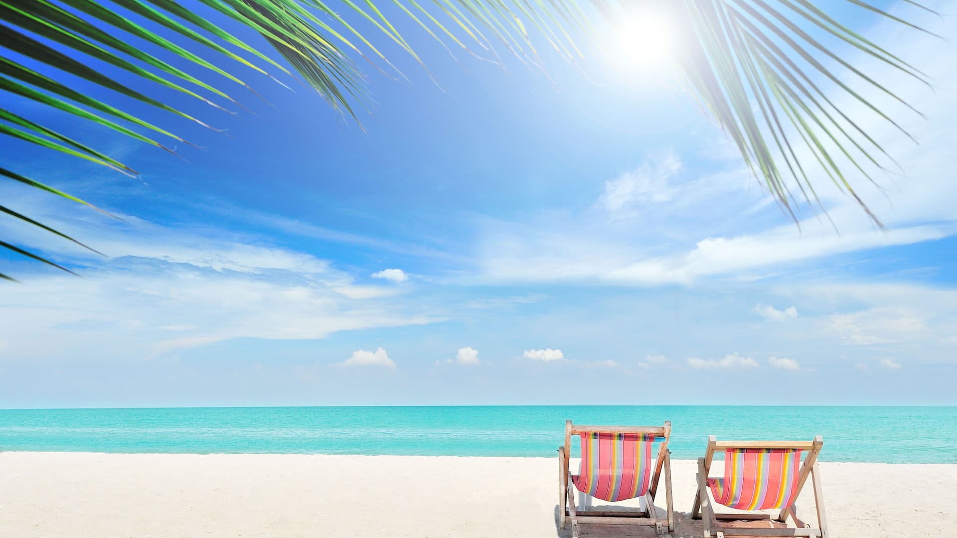 Two colorful striped beach chairs sit on the white sand beach of Great Stirrup Cay, Bahamas, facing the clear turquoise water under a bright blue sky with palm fronds overhead.