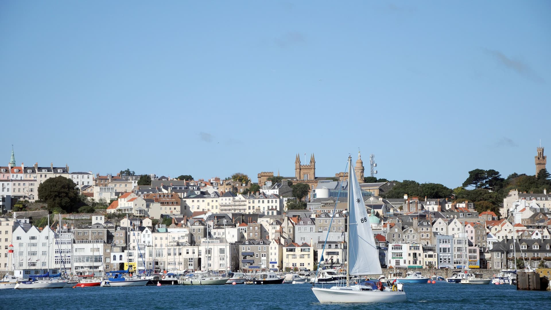 A sailboat gracefully sailing in the harbor of St. Peter Port, Guernsey, with the town's historic buildings and spires sprawling up the hillside in the background.