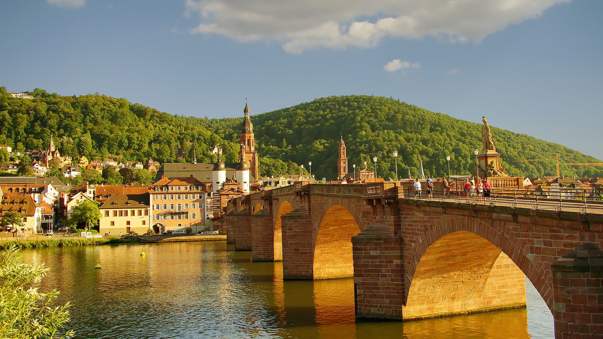 A picturesque view of Heidelberg, Germany, with the historic Old Bridge spanning the Neckar River, a town with colorful buildings on the bank, and lush green hills in the background.
