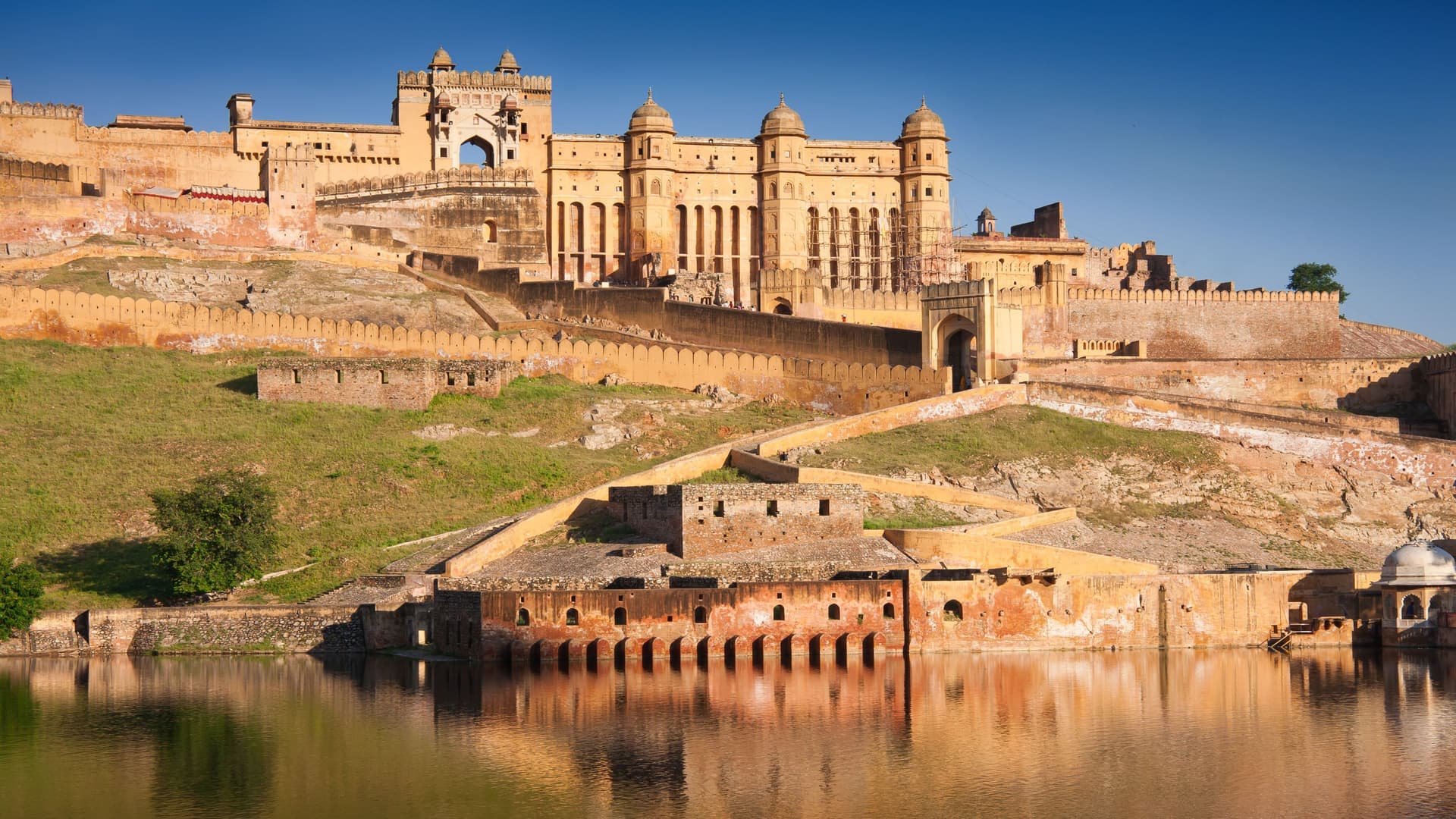 A wide-angle view of the majestic Amber Fort and Palace in Jaipur, India, with its ornate walls and towers reflecting in the serene Maota Lake.