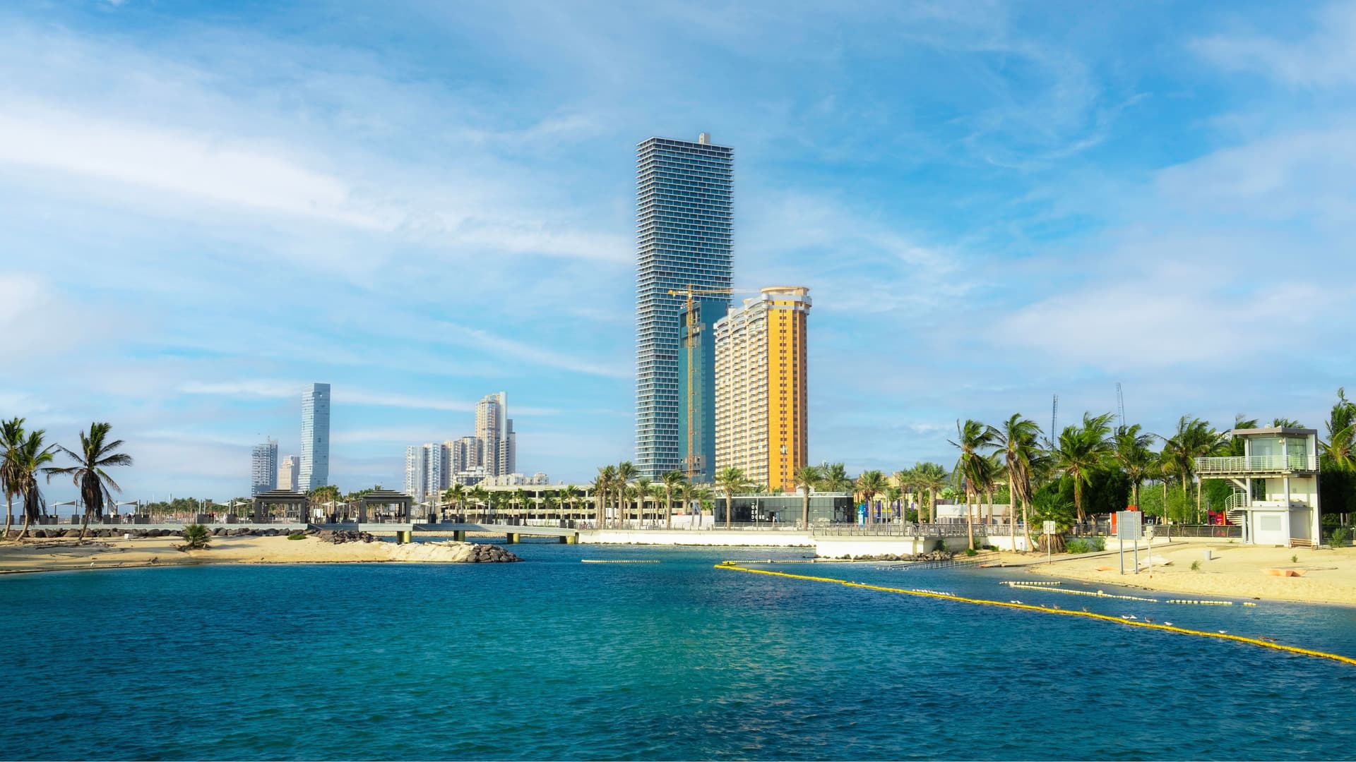 A scenic view of Jeddah's modern skyline, with towering skyscrapers and lush palm trees lining a sandy beach and a clear blue bay.