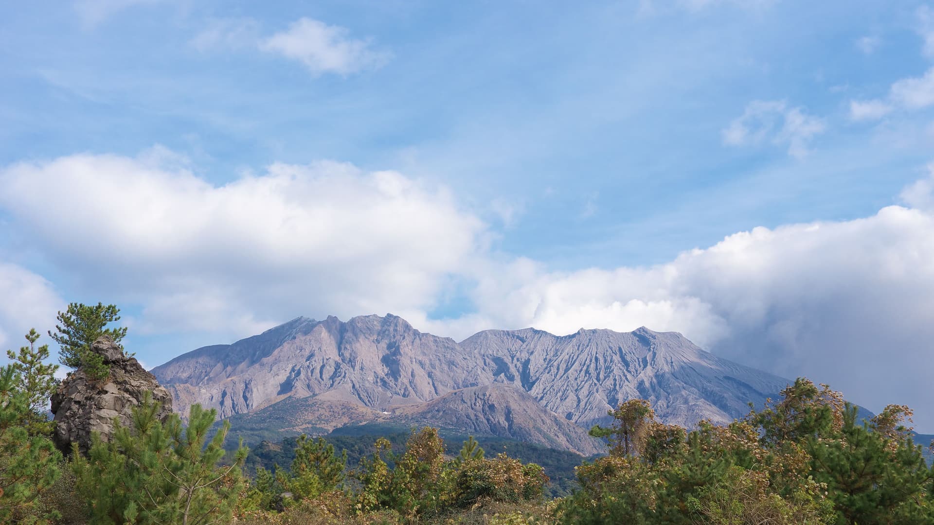 A striking view of the active Sakurajima volcano and its peaks, rising behind a foreground of green trees, in the bay of Kagoshima, Japan.