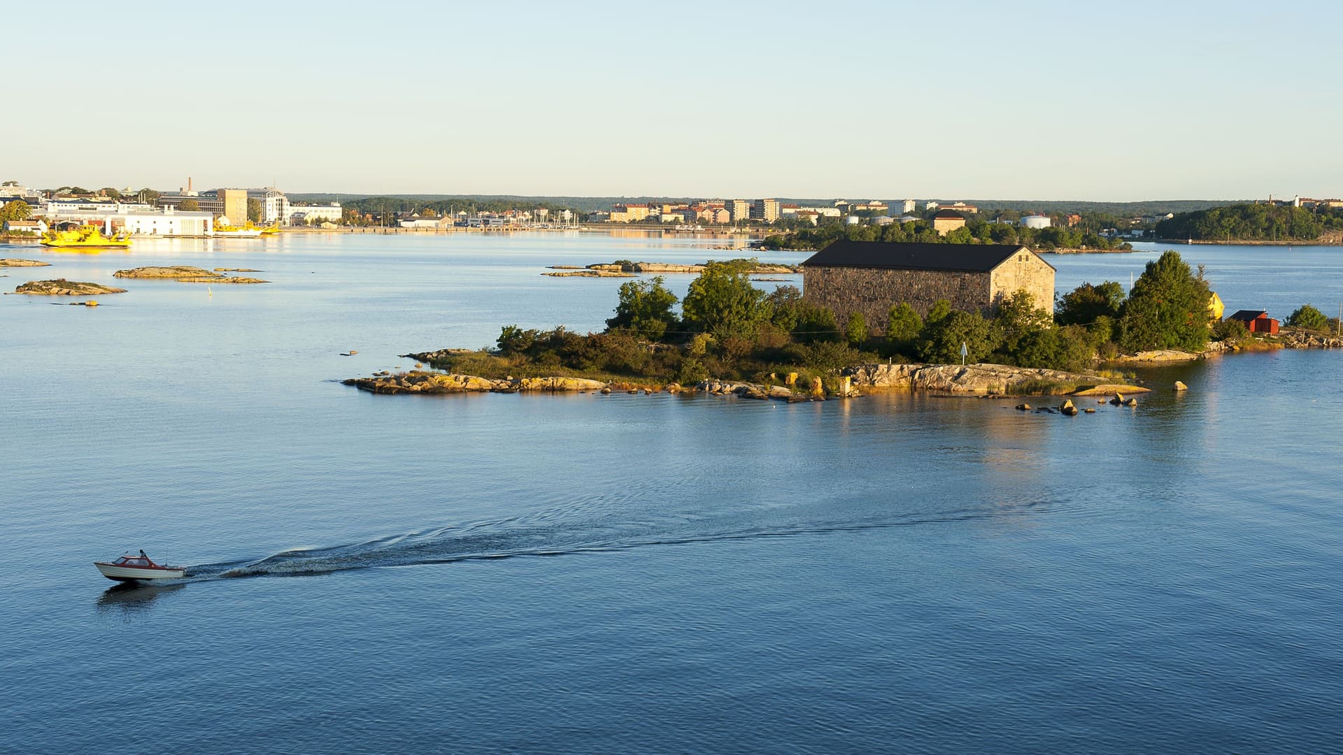 A tranquil landscape of the Karlskrona archipelago in Sweden, featuring a small island with a historic stone building and trees in the foreground, and the modern city skyline across the calm Baltic Sea in the distance.