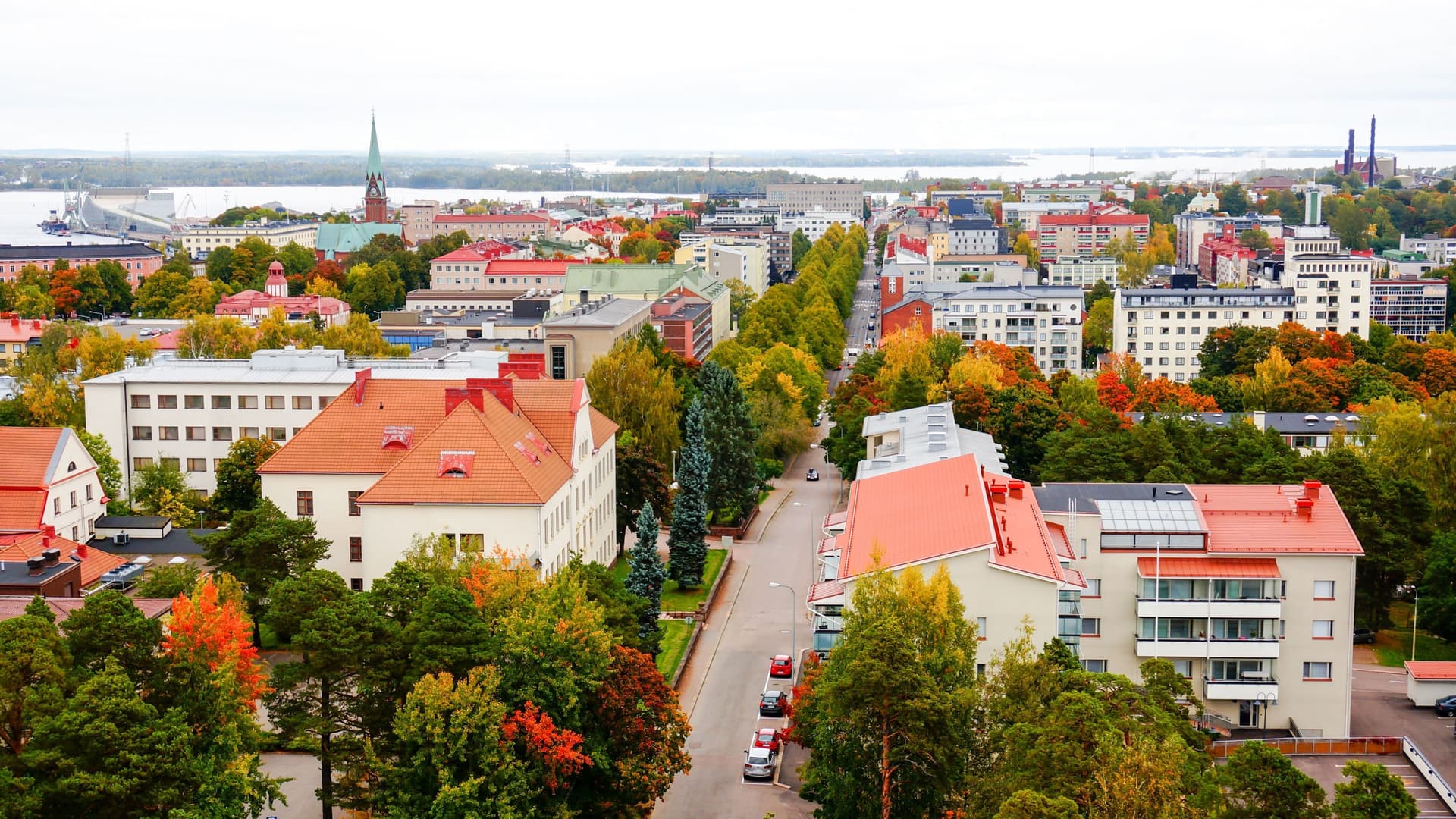 An aerial view of the city of Kotka in Finland, showcasing a colorful autumn cityscape with a variety of buildings and trees along a wide street, leading to the harbor in the distance.