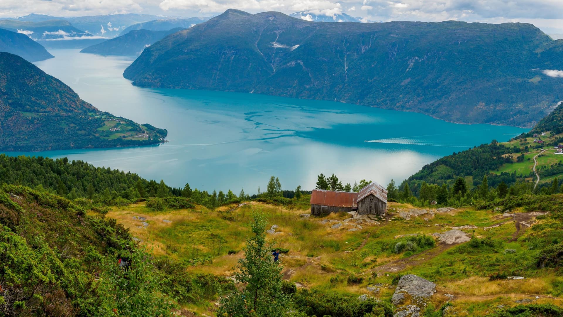 A breathtaking panoramic shot of the Lustrafjord in Norway, showcasing the turquoise waters, lush green landscape with a rustic hut, and majestic mountains under a cloudy sky.