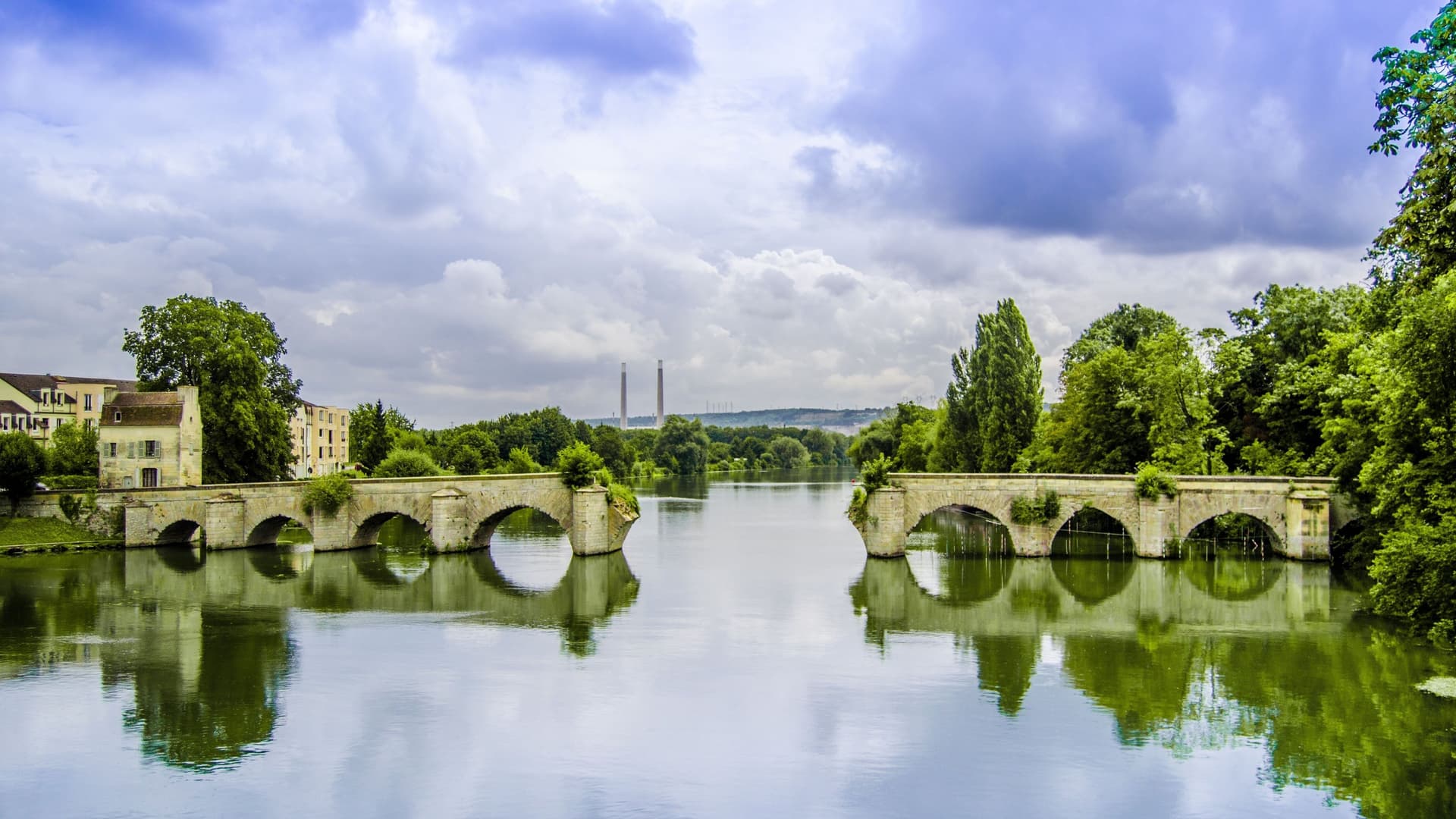 A picturesque view of the old stone bridge with multiple arches spanning the calm waters of the Seine River in Mantes-la-Jolie, France.