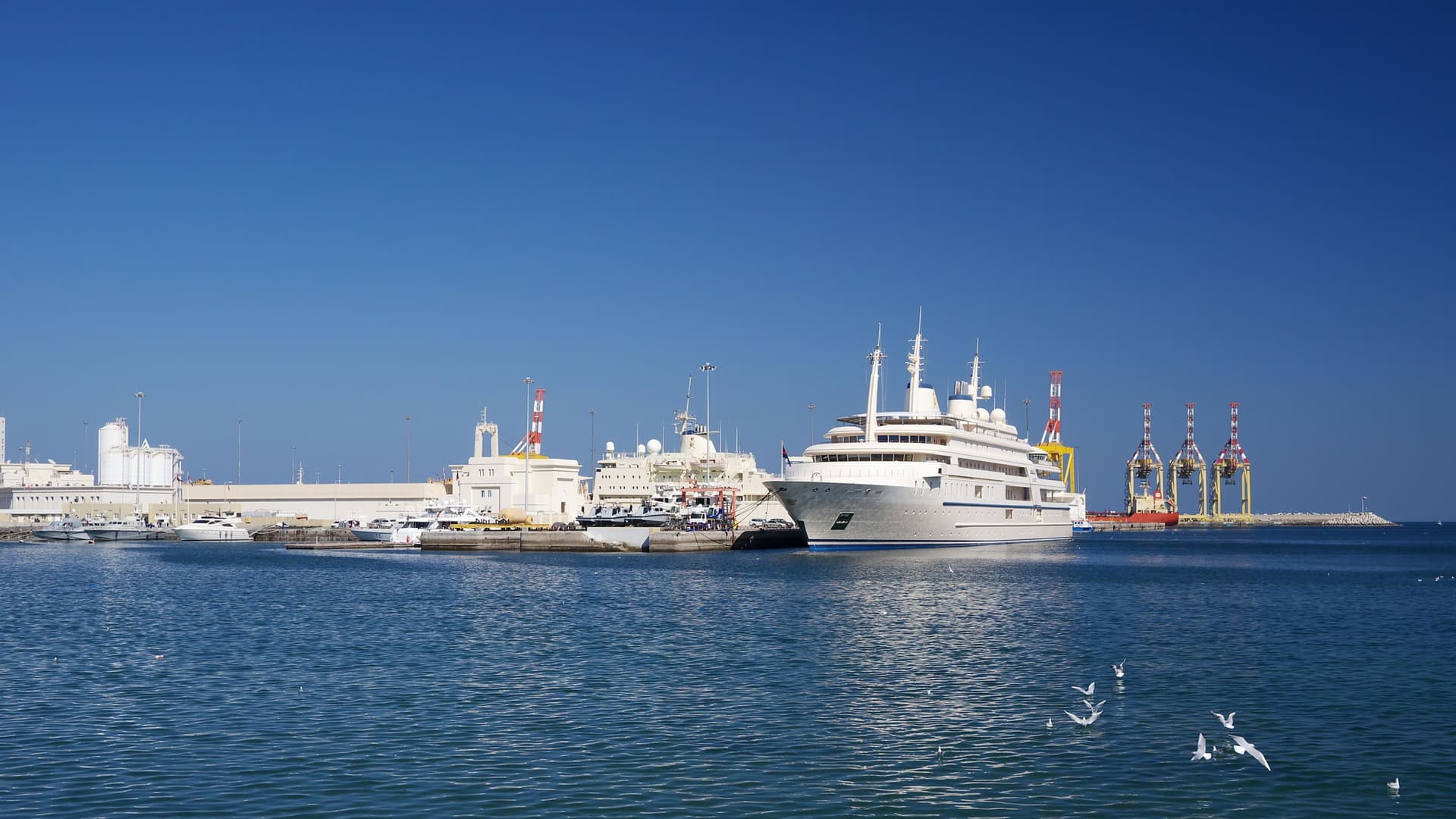 A high-angle view of Port Sultan Qaboos in Muscat, Oman, with a large cruise ship or cargo vessel docked in the harbor. The port is backed by the residential buildings of the city's waterfront, which climb the surrounding rugged, arid mountains. The scene captures the commercial and scenic beauty of Muscat's main port.