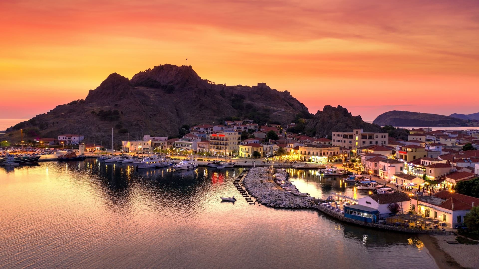 A vibrant sunset view of the harbor and town of Myrina, Greece, with boats docked and lights illuminating the buildings against a backdrop of a mountain with a castle ruin.