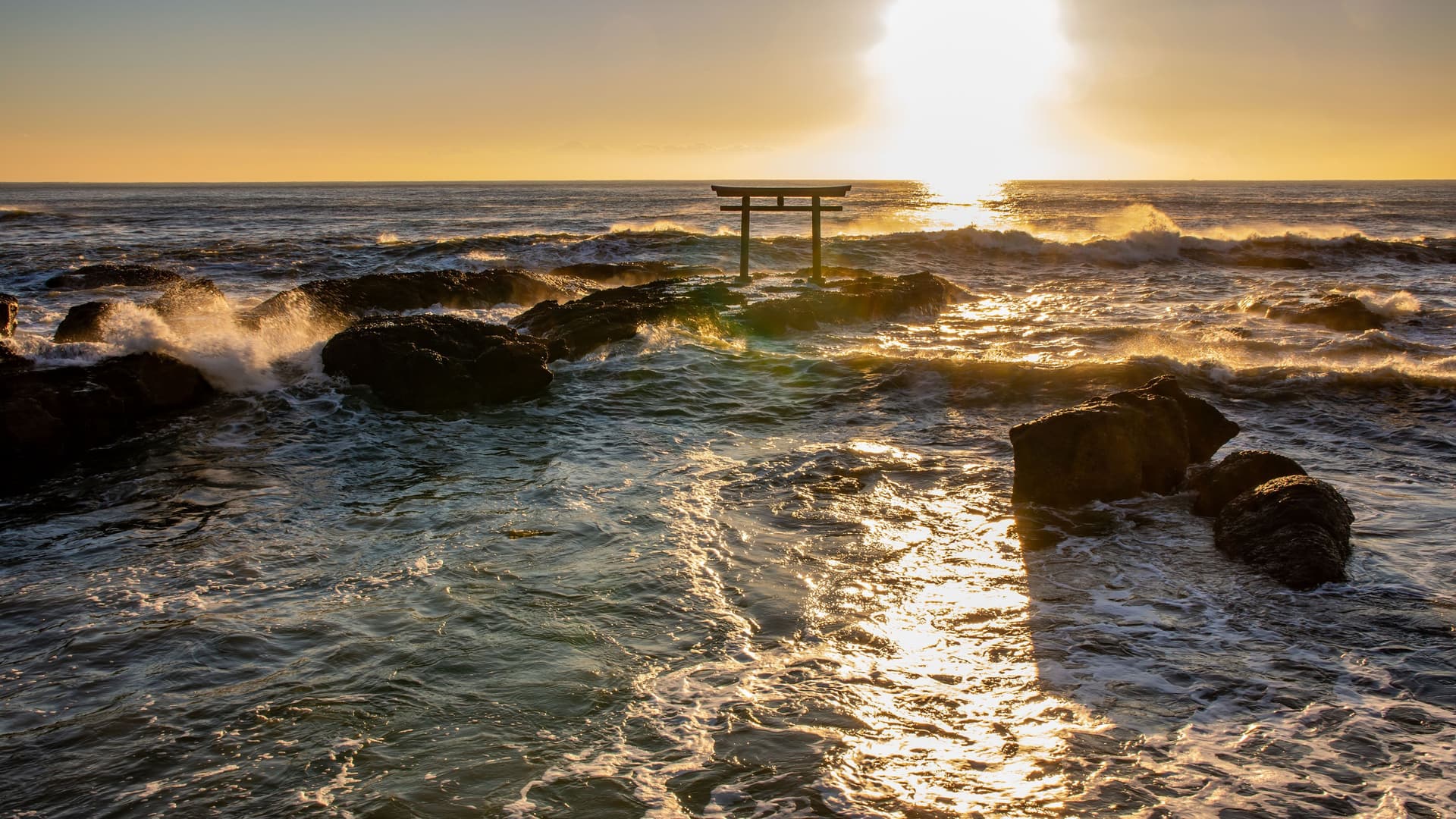 A beautiful sunrise over the Pacific Ocean, with the iconic torii gate of the Oarai Isosaki Shrine in Japan standing on a rocky outcrop amidst the splashing waves.