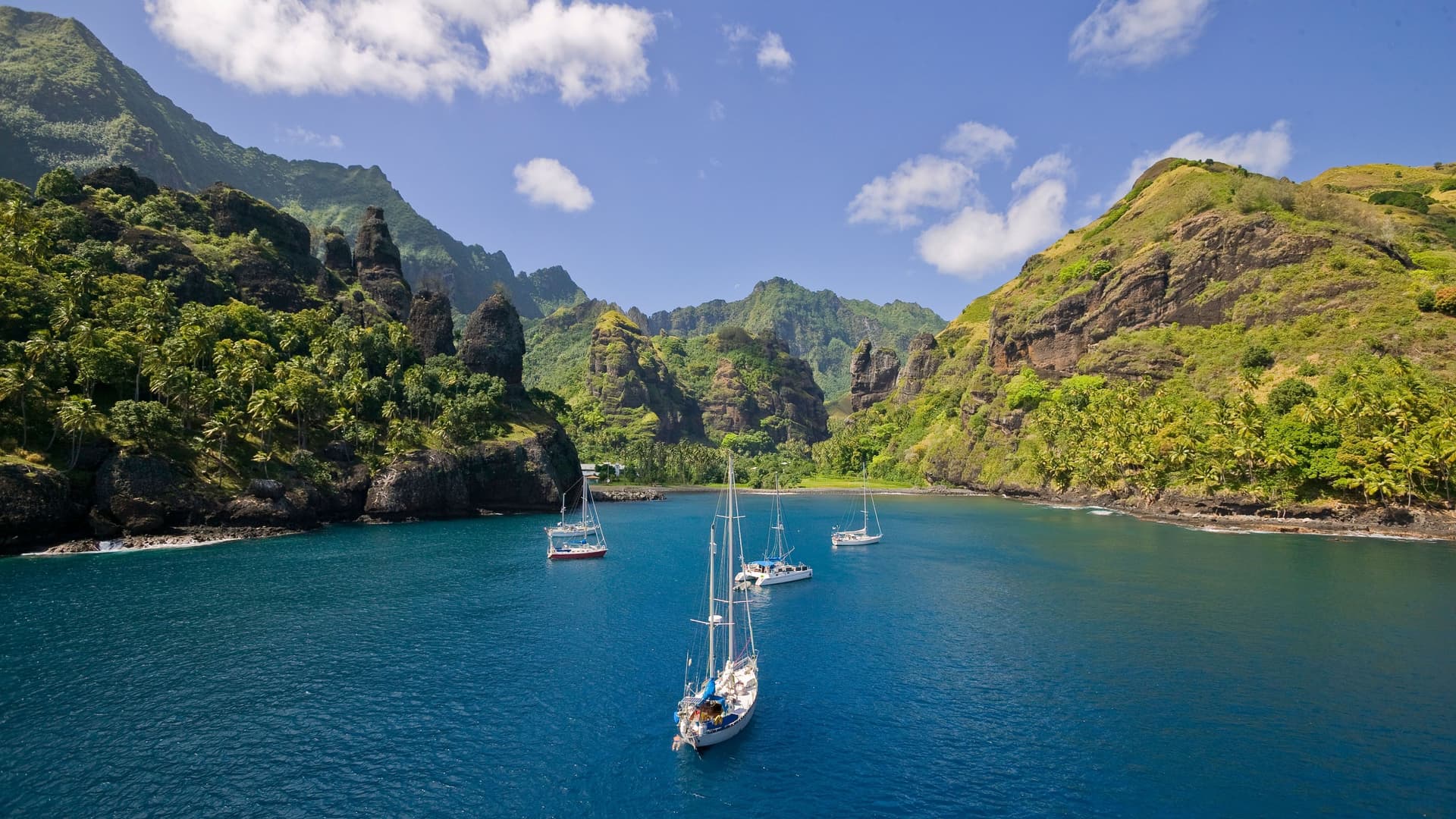 A beautiful scenic view of Omoa Bay on Fatu Hiva, Marquesas Islands, with several sailboats anchored in the peaceful blue water, surrounded by dramatic green cliffs and lush tropical vegetation.