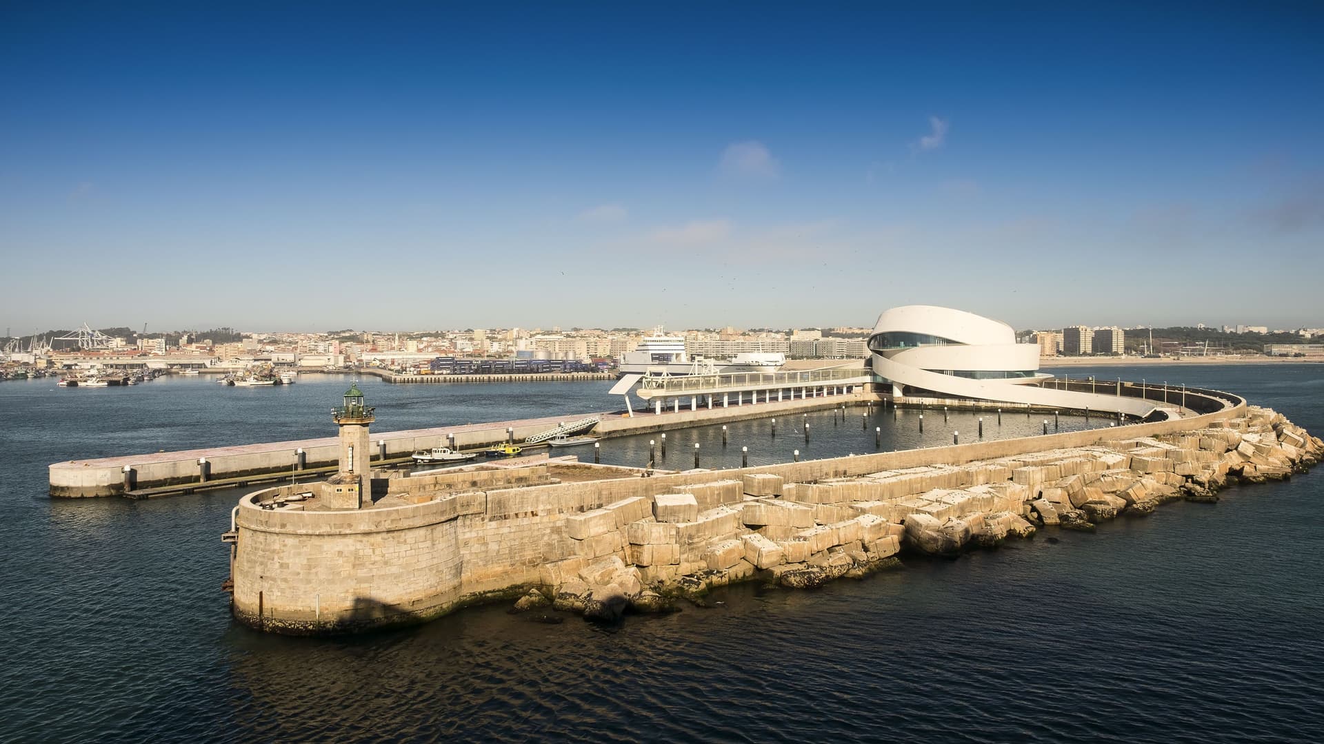 A high-angle view of the modern, white spiral-shaped cruise terminal at the Port of Leixões, Oporto, Portugal, extending into the ocean next to a rocky breakwater.