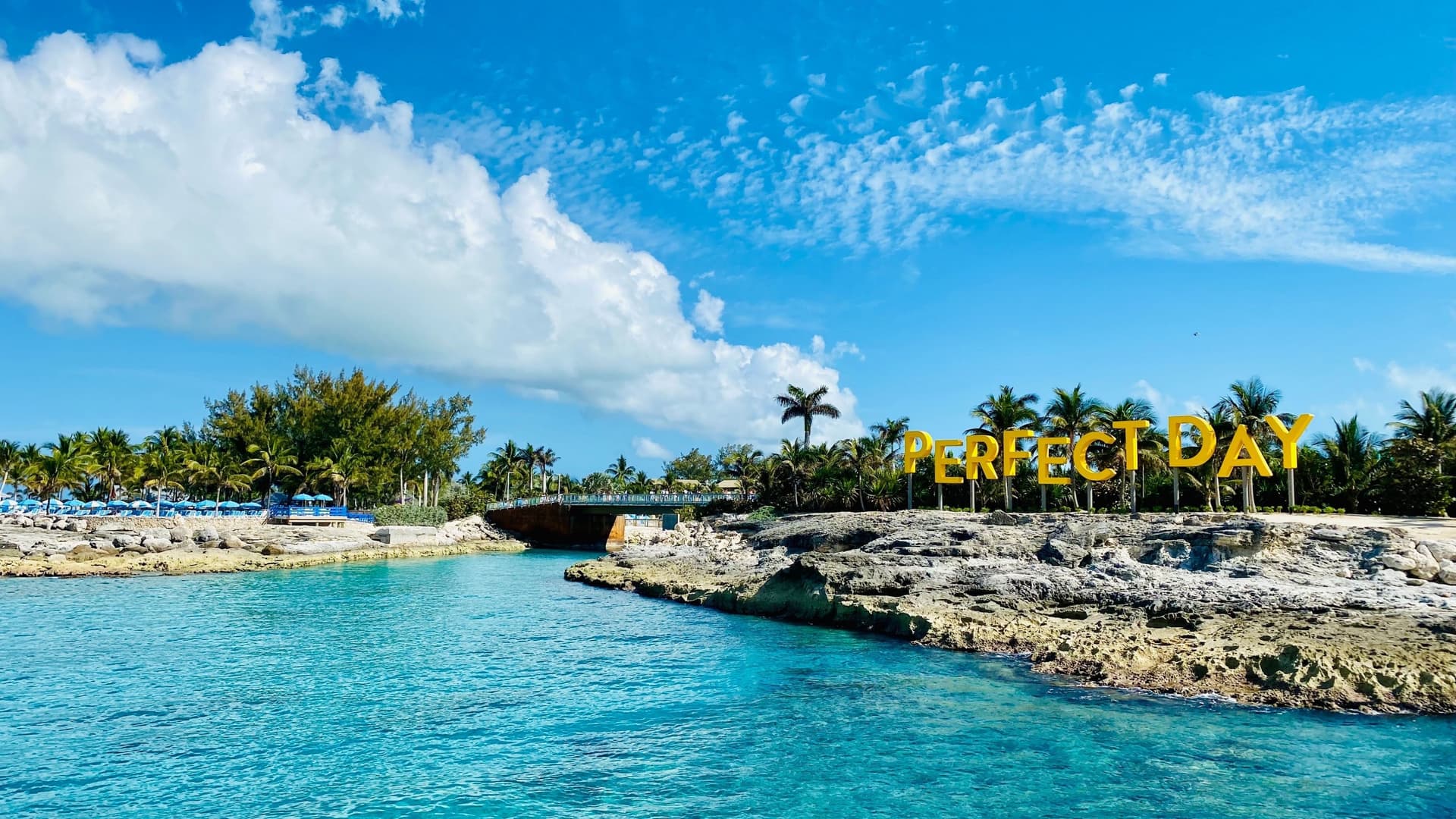 A view of a huge sign that reads "Perfect Day at CocoCay," a private island in the Bahamas, with turquoise water and palm trees under a bright blue sky.