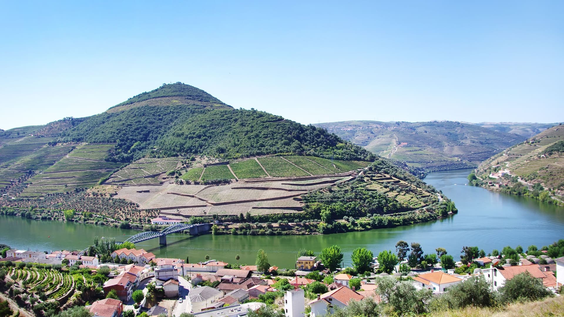 A panoramic view of the Douro River in Portugal, showcasing the terraced vineyards on the hillsides surrounding the small town of Pinhão, with a bridge crossing the water.