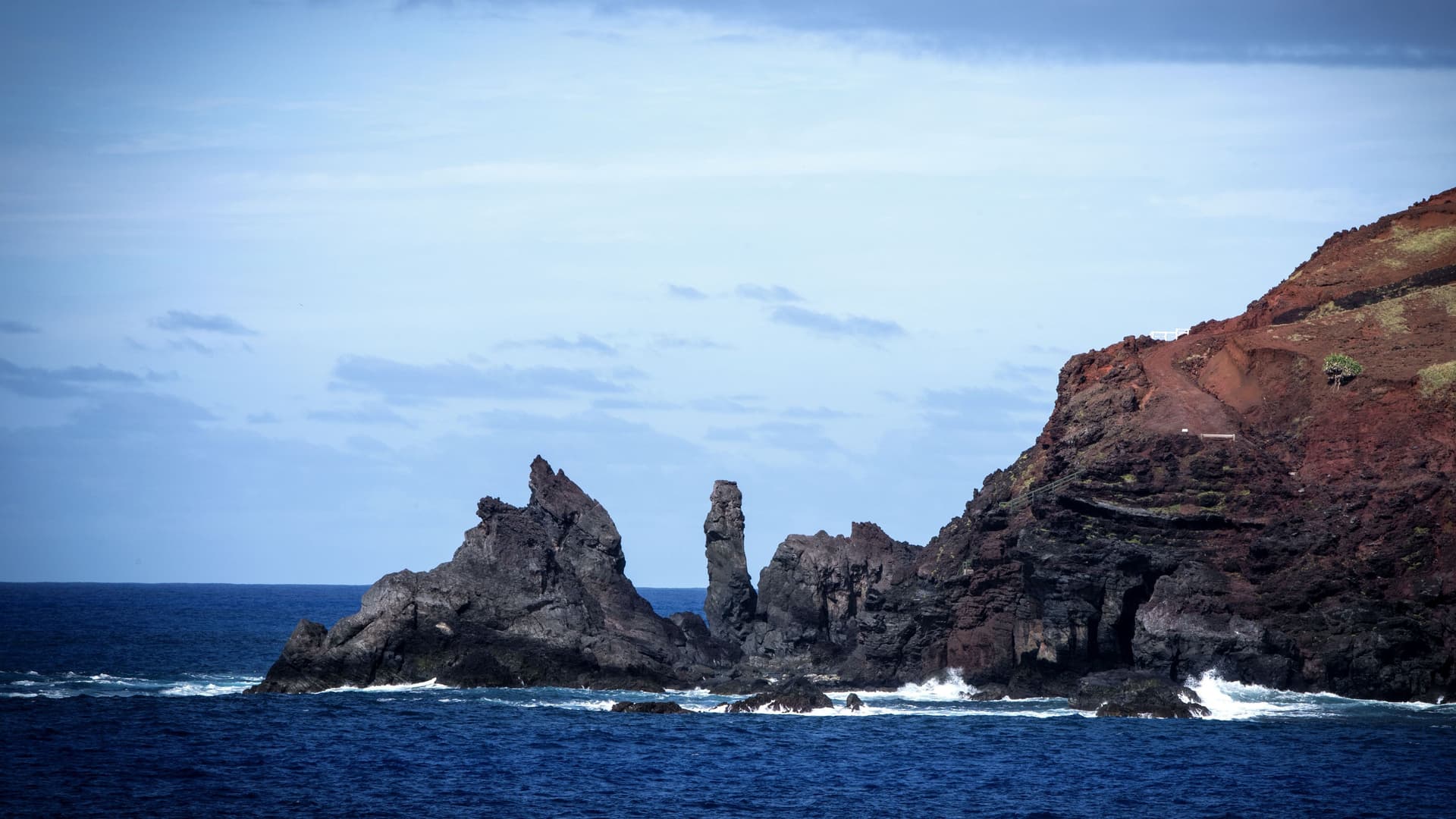 A scenic view of the rugged, volcanic coastline of Pitcairn Island in the South Pacific, with towering dark cliffs and unique rock formations jutting out of the deep blue water. 