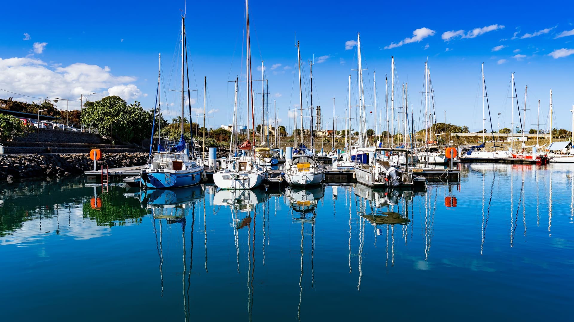 A scenic view of the marina at Pointe des Galets on Réunion Island, showing sailboats and yachts docked in the calm blue water with their reflections visible, under a clear sky.