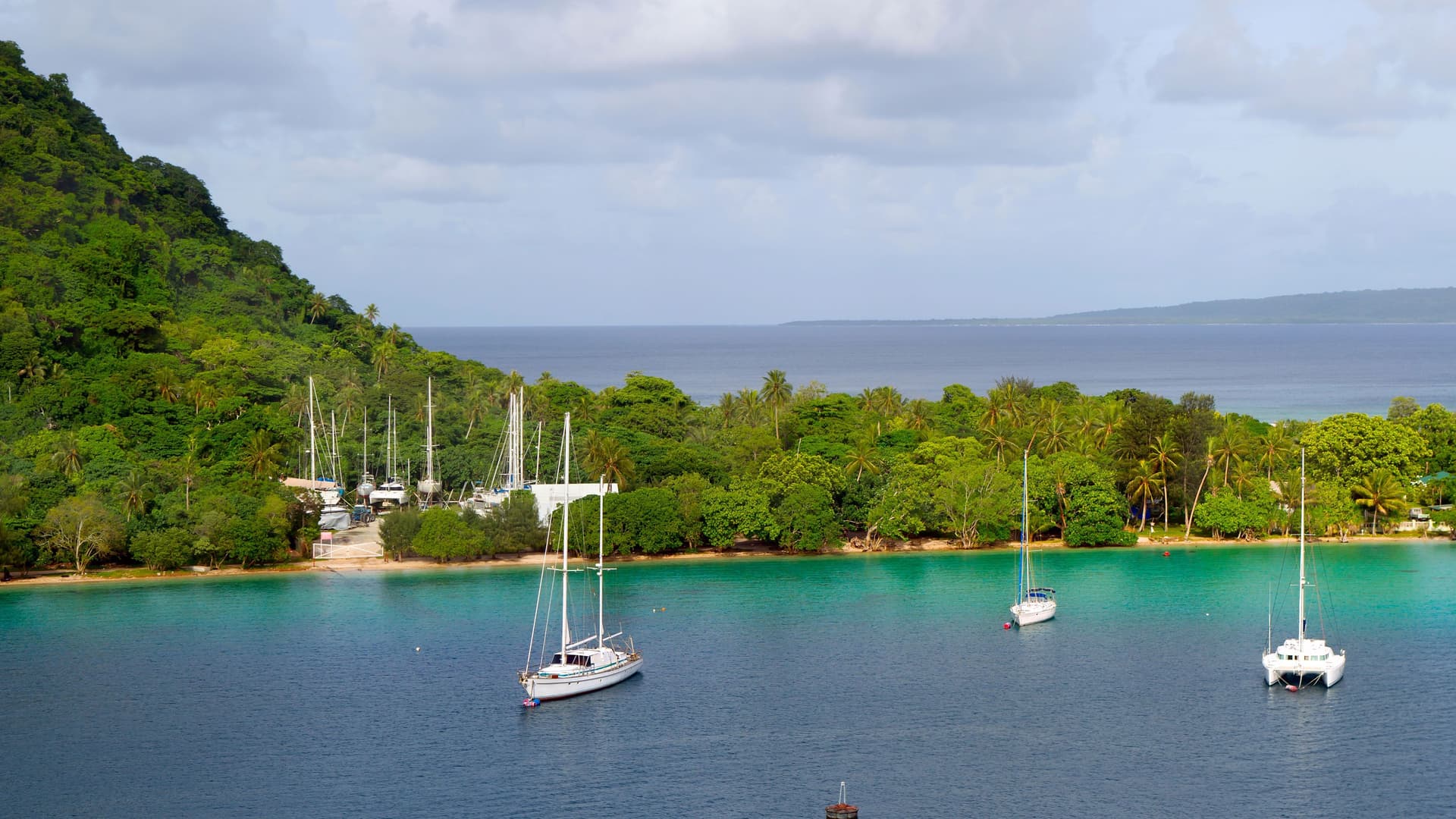 A beautiful tropical bay in Port Vila, Vanuatu, with several sailboats and yachts anchored in the clear turquoise water, surrounded by lush green trees on the coastline.