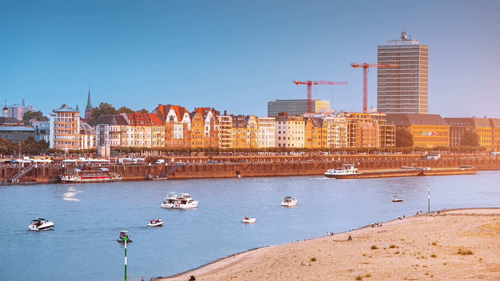 The Rhine River waterfront features boats, a sandy bank in the foreground, and historic, colorful buildings in the sunshine.