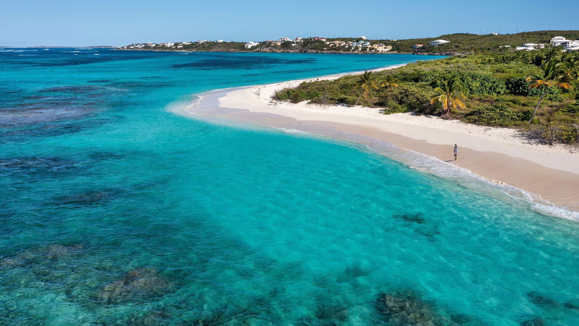 An aerial view of Road Bay, Anguilla, showing a pristine white sand beach, lush green vegetation, and the stunningly clear turquoise water of the Caribbean Sea.