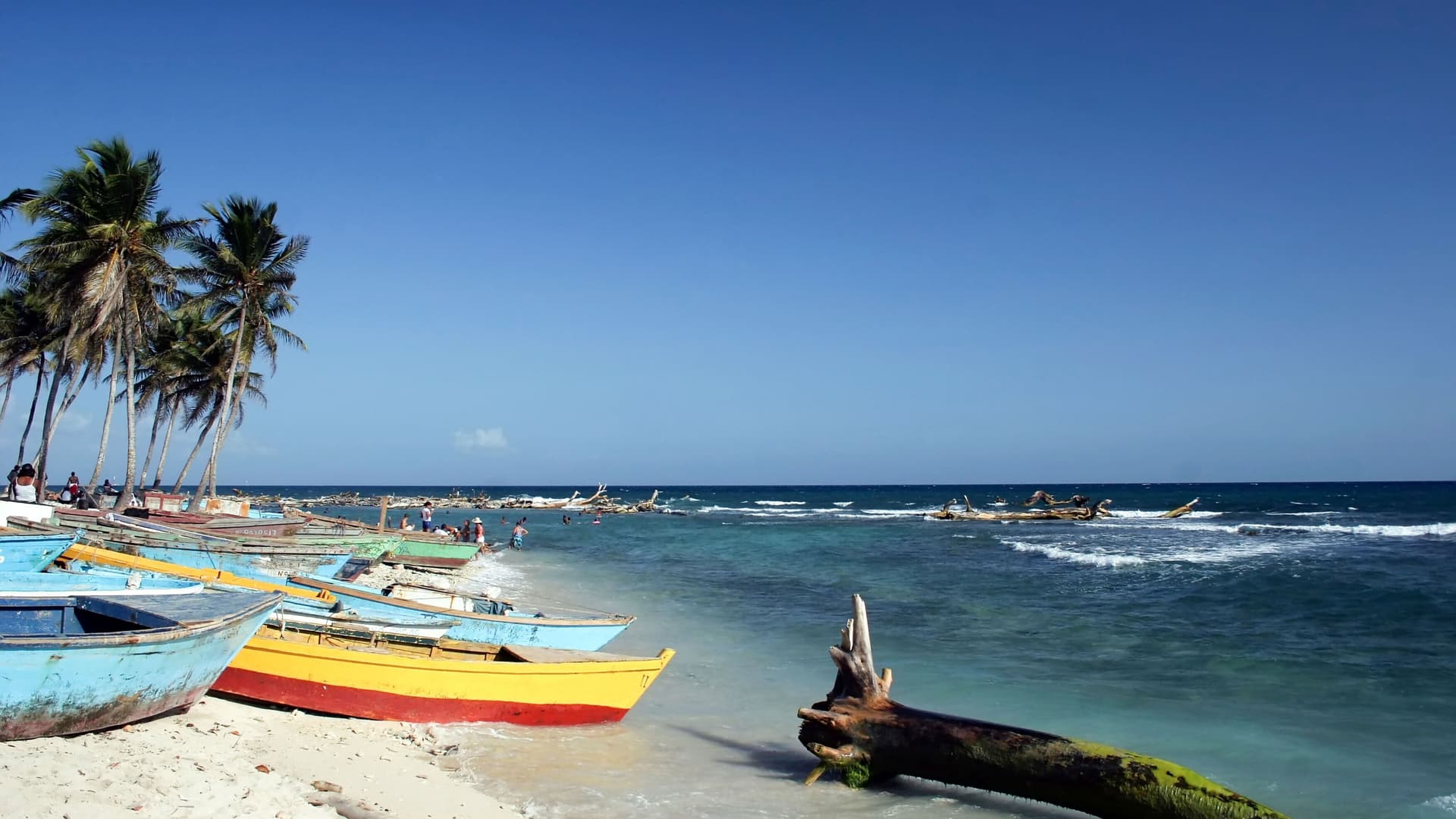 This is a scenic image of a tropical beach in Santo Domingo, with multiple colorful fishing boats resting on the sandy shore, tall palm trees, and the calm turquoise sea under a bright, clear sky.