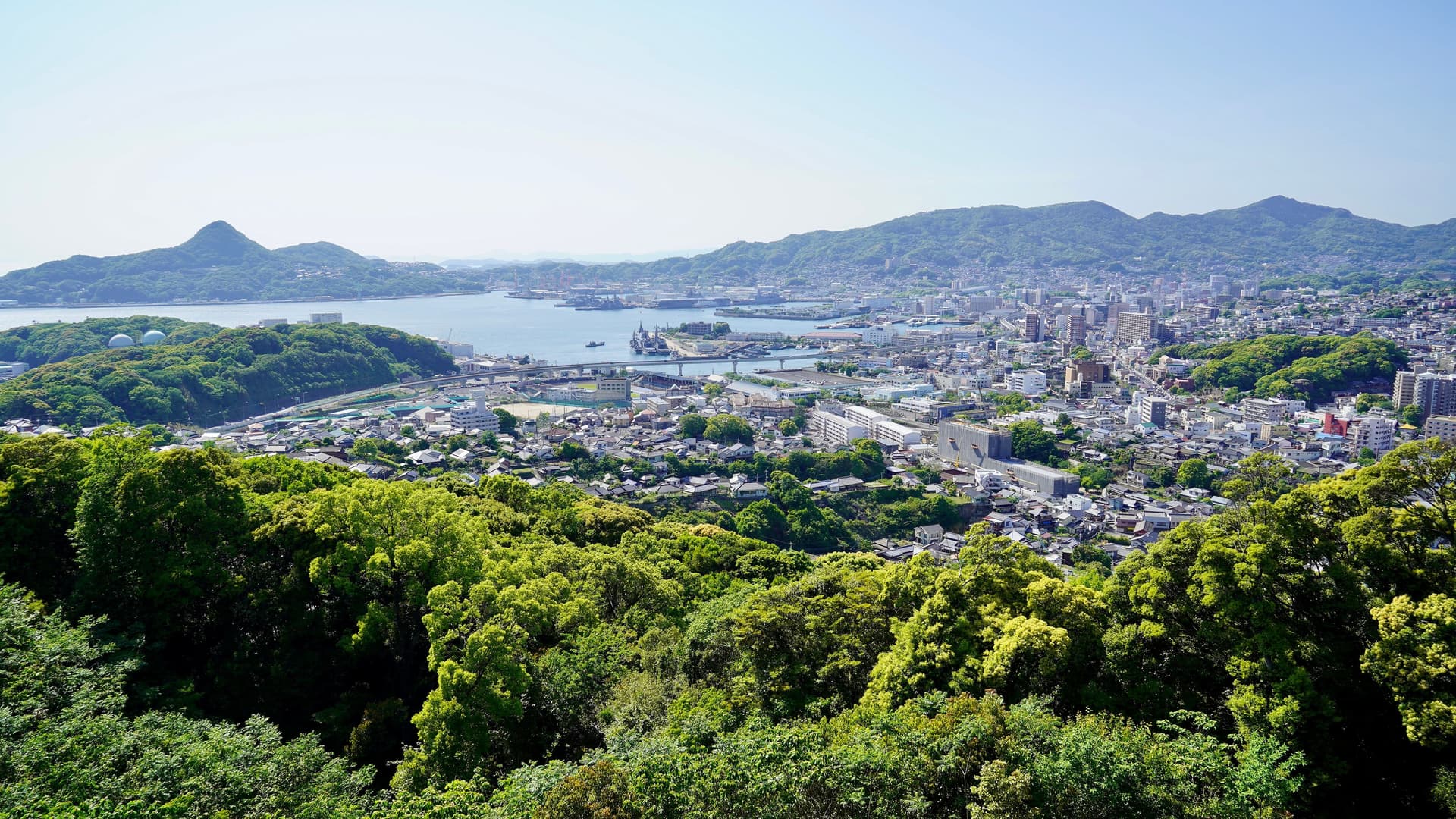 A scenic high-angle view of the port city of Sasebo in Nagasaki, Japan, nestled between lush green forests and the serene blue bay.