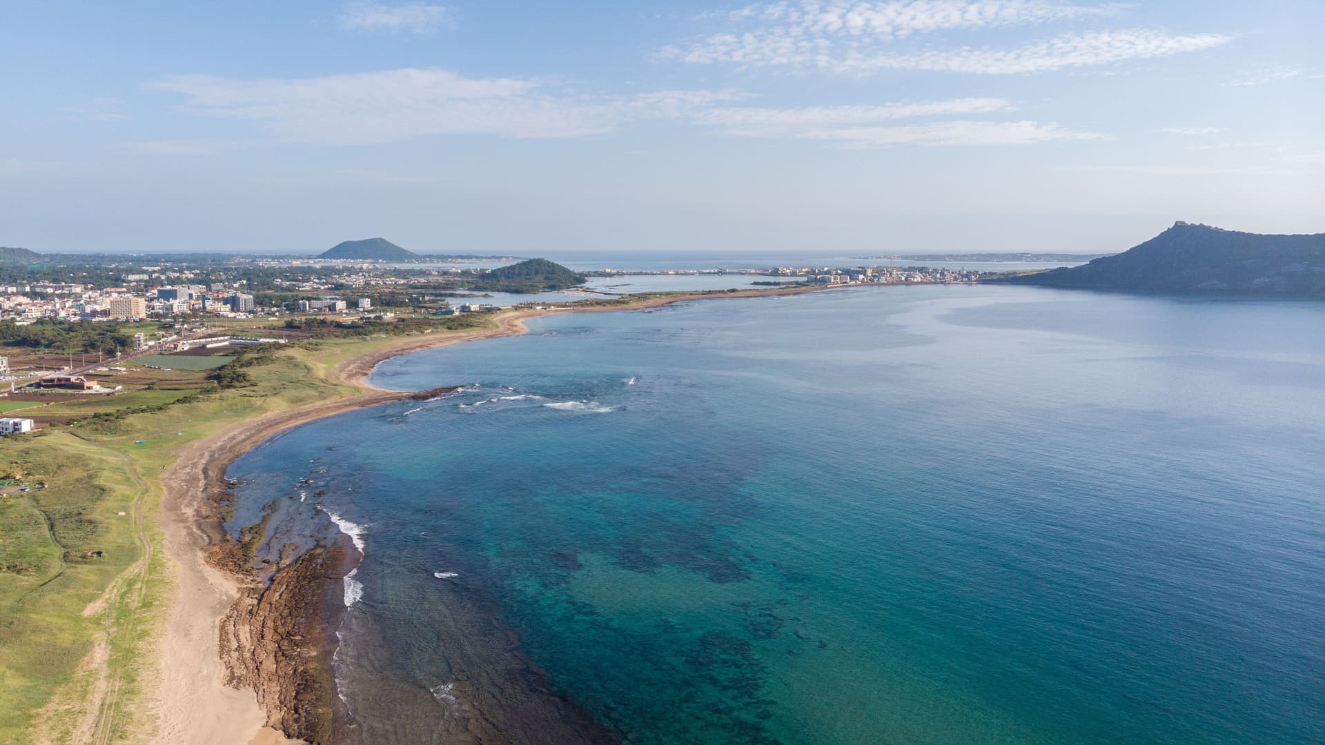 An aerial view of the scenic coastline of Seogwipo City, South Korea, with a curving beach, rocky shores, and the tranquil sea against a backdrop of mountains and the city skyline.
