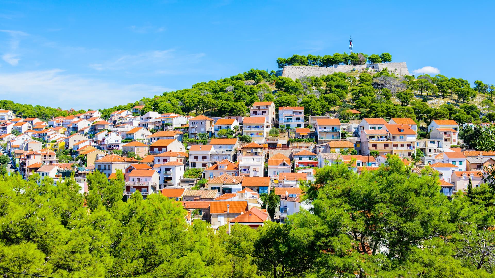 A panoramic daytime shot of the historic city of Šibenik, Croatia, with its red-roofed buildings climbing a hill to the hilltop fortress.