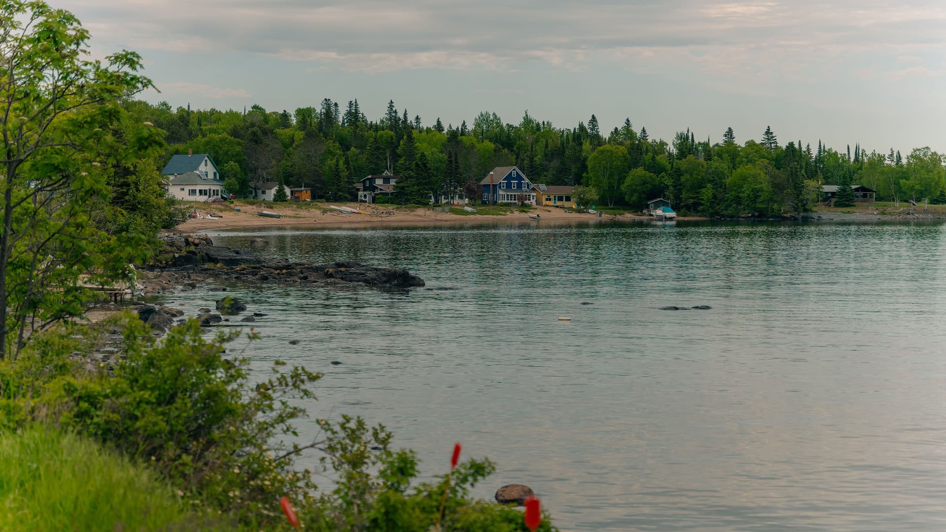 A tranquil view of Silver Islet, Canada, with a few colorful cottages and docks along the rocky shoreline of Lake Superior, surrounded by lush green trees and a quiet sky.