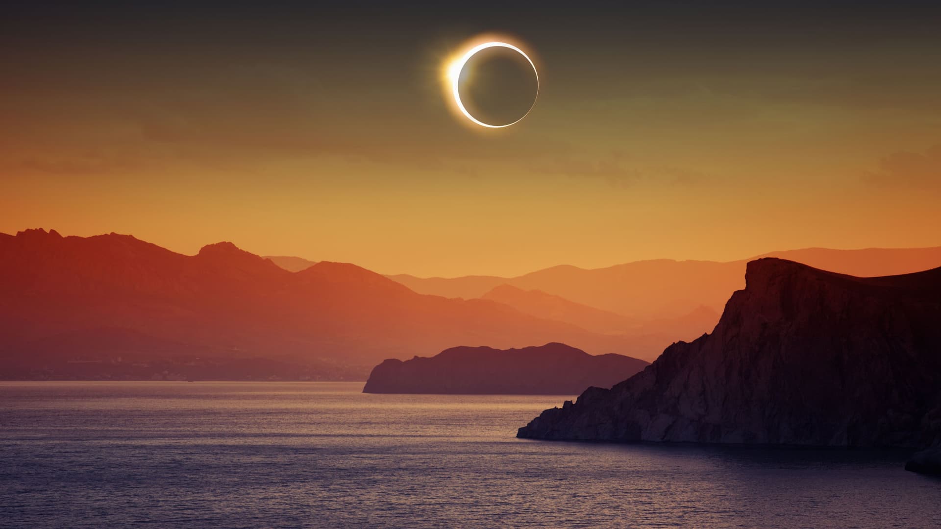 A dramatic celestial event over the sea with a ring of light, the total solar eclipse, visible over a tranquil body of water surrounded by silhouetted mountains.
