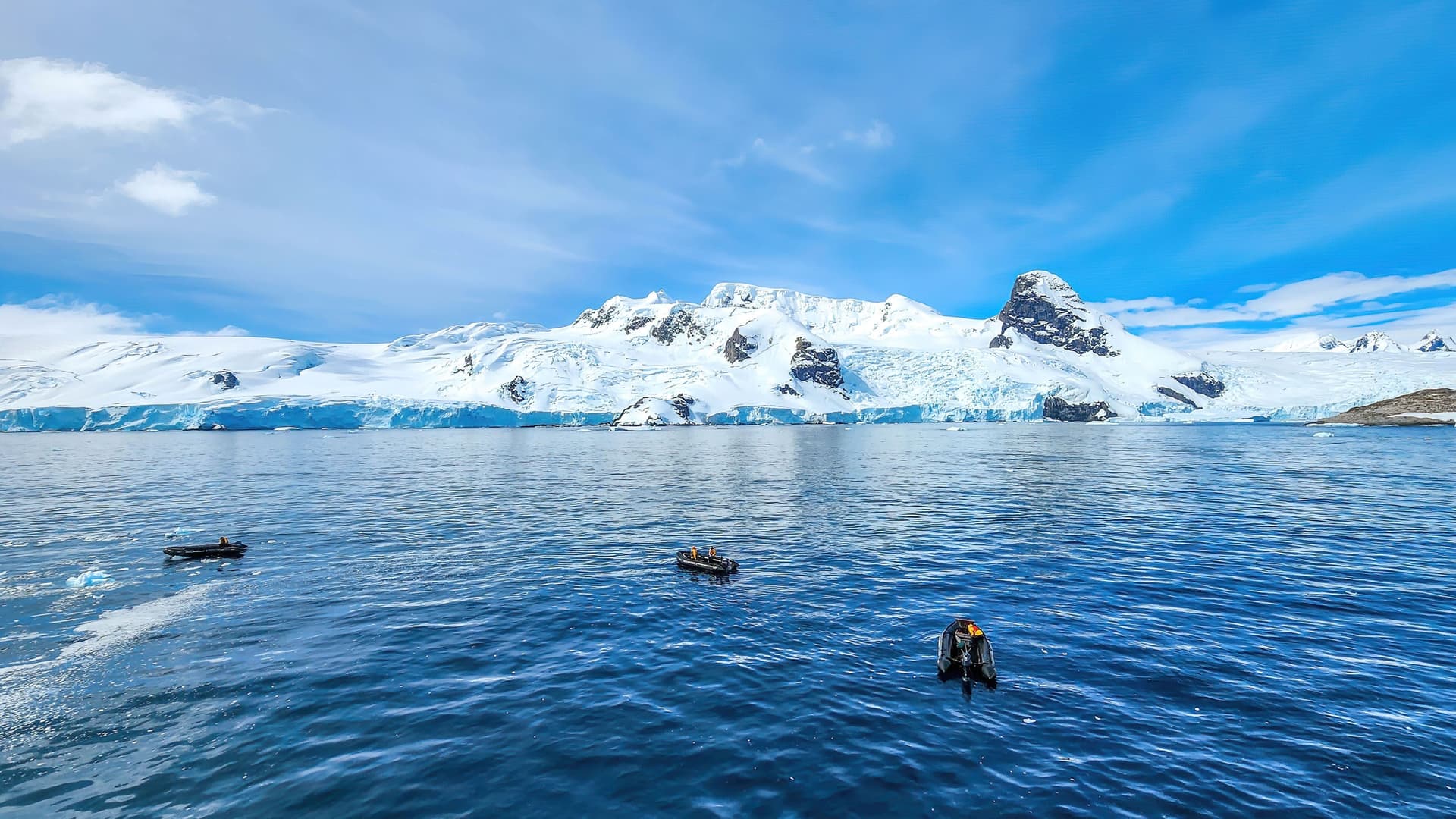 A stunning panoramic view of the South Shetland Islands, with a majestic snow-covered coastline and glaciers meeting the deep blue water, with a few small boats in the foreground.