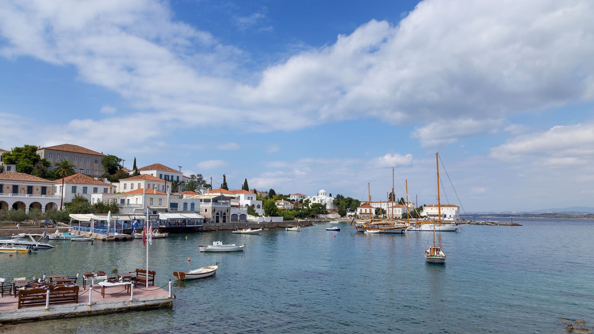 A charming view of the old harbor in Spetses, Greece, with traditional white buildings and classic wooden sailboats moored in the calm bay under a bright sky with scattered clouds.