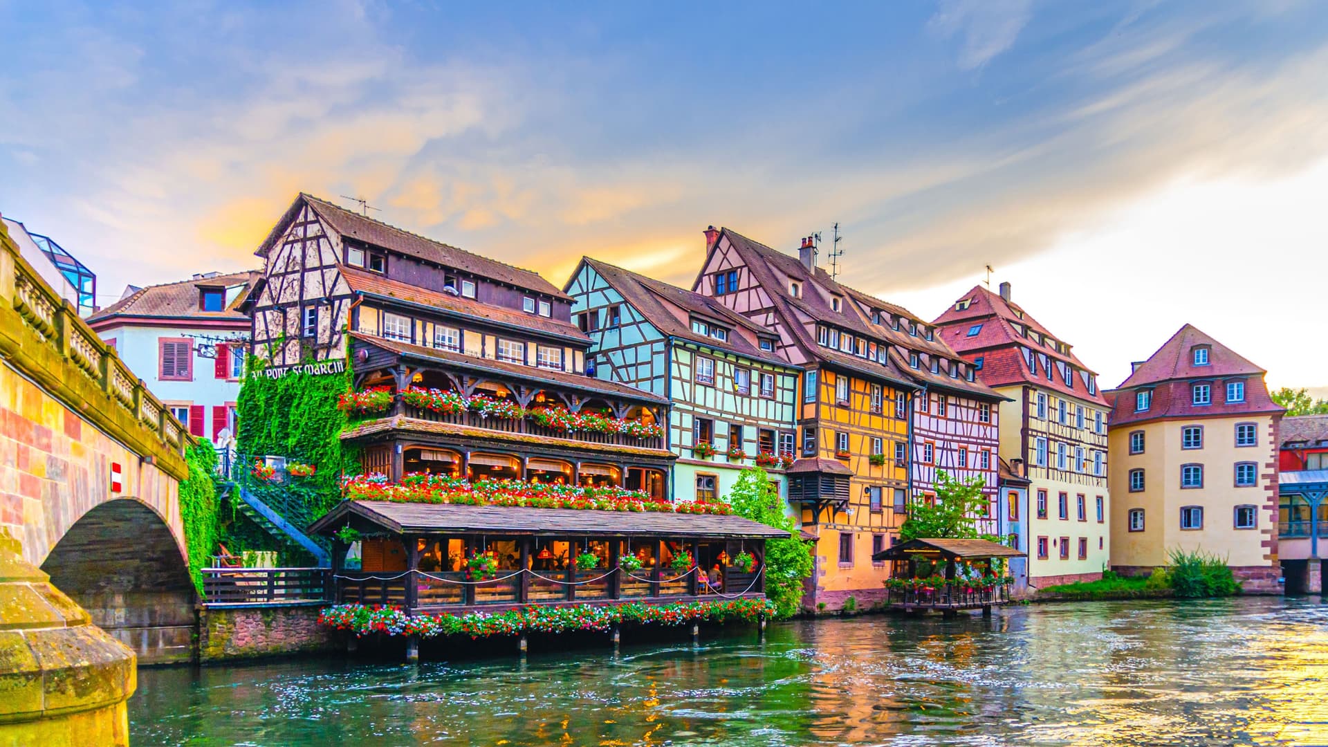 A wide-angle view of the colorful half-timbered buildings in Petite France, Strasbourg, with a bridge over the Ill River and flowers hanging from the balconies.