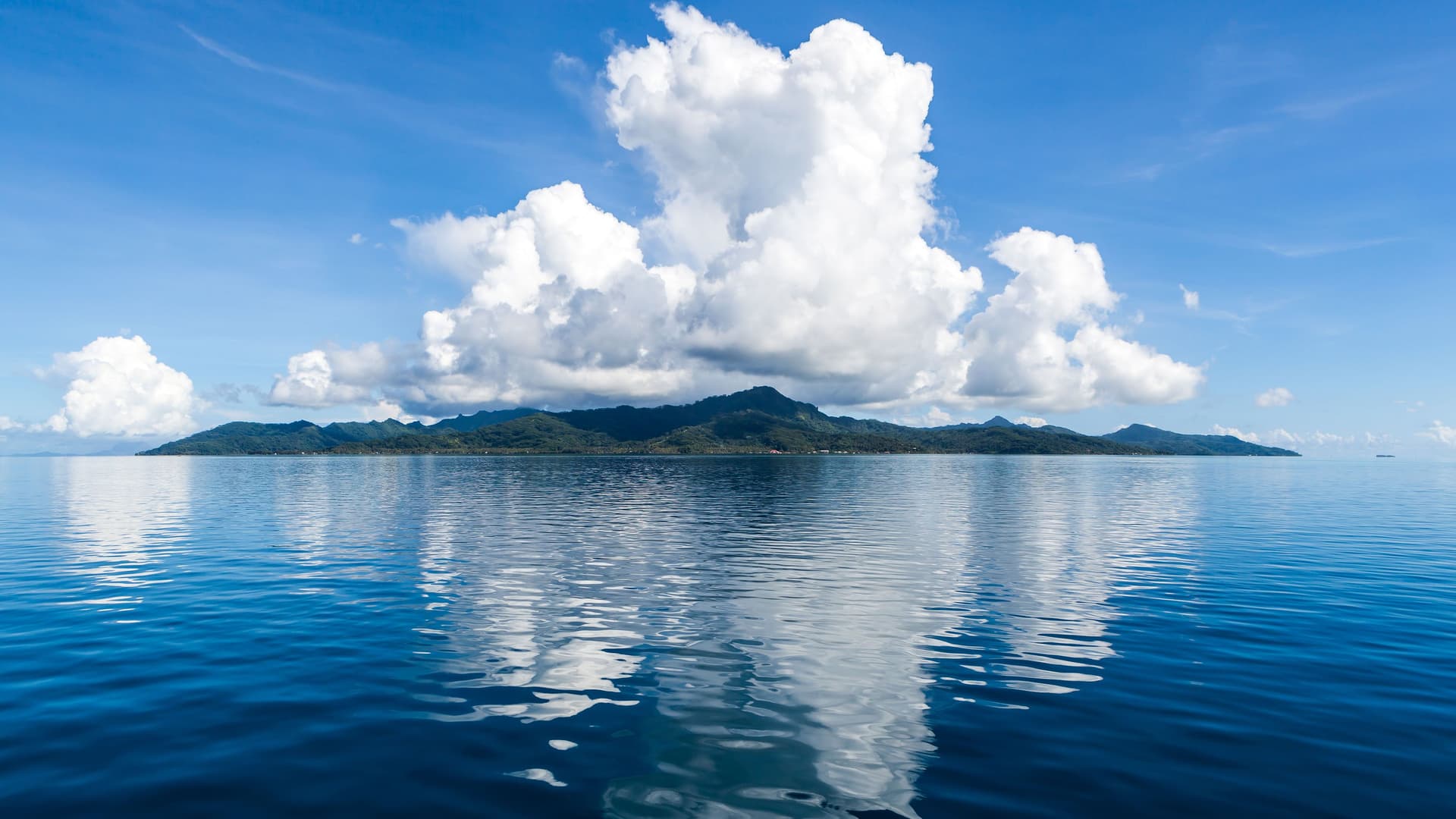 A beautiful panoramic view of the island of Taha'a, French Polynesia, with lush green hills and a large, puffy white cloud formation reflecting in the calm, deep blue water under a clear blue sky.