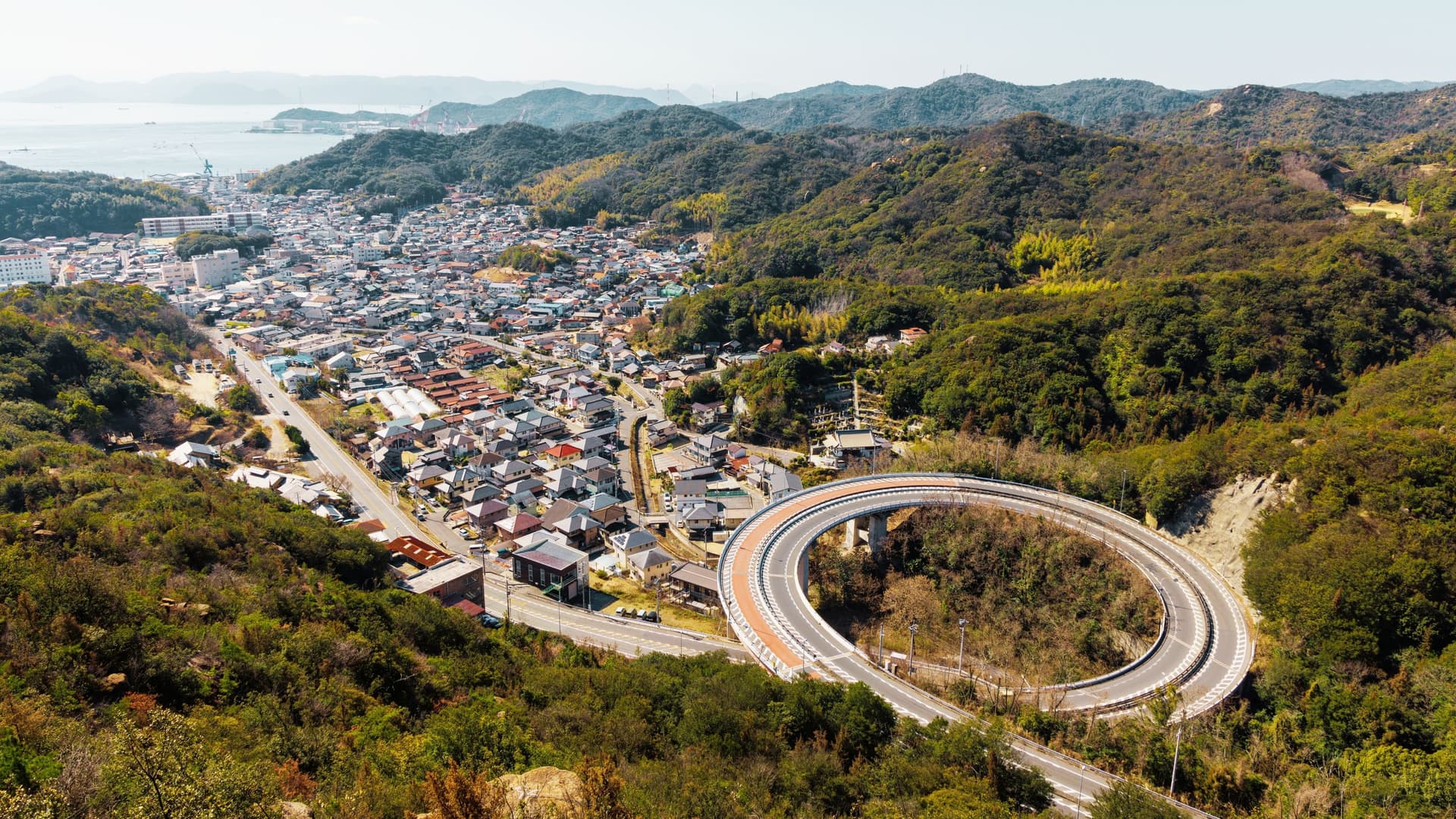 An aerial view of a large loop bridge (also known as a spiral bridge or helical bridge) in Tamano City, Okayama Prefecture, Japan. The elevated highway winds down in a complete circle to efficiently manage the change in elevation, connecting a high coastal road or mountainous area to the lower terrain below. The surrounding landscape features lush green trees and forested hillsides.