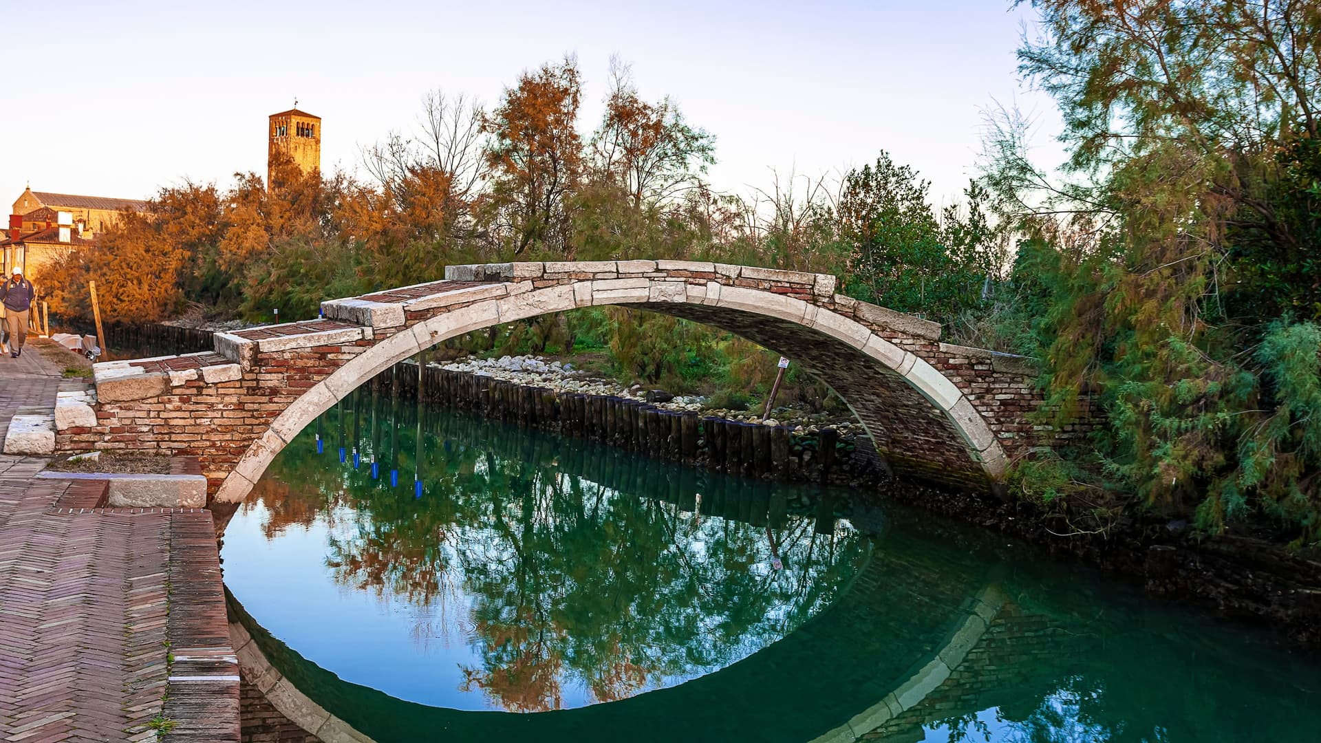 A peaceful canal scene in Torcello with a stone arch bridge forming a near‑perfect circle in its reflection, a cobblestone path where two people walk, and autumn trees and a historic bell tower glowing in warm light.