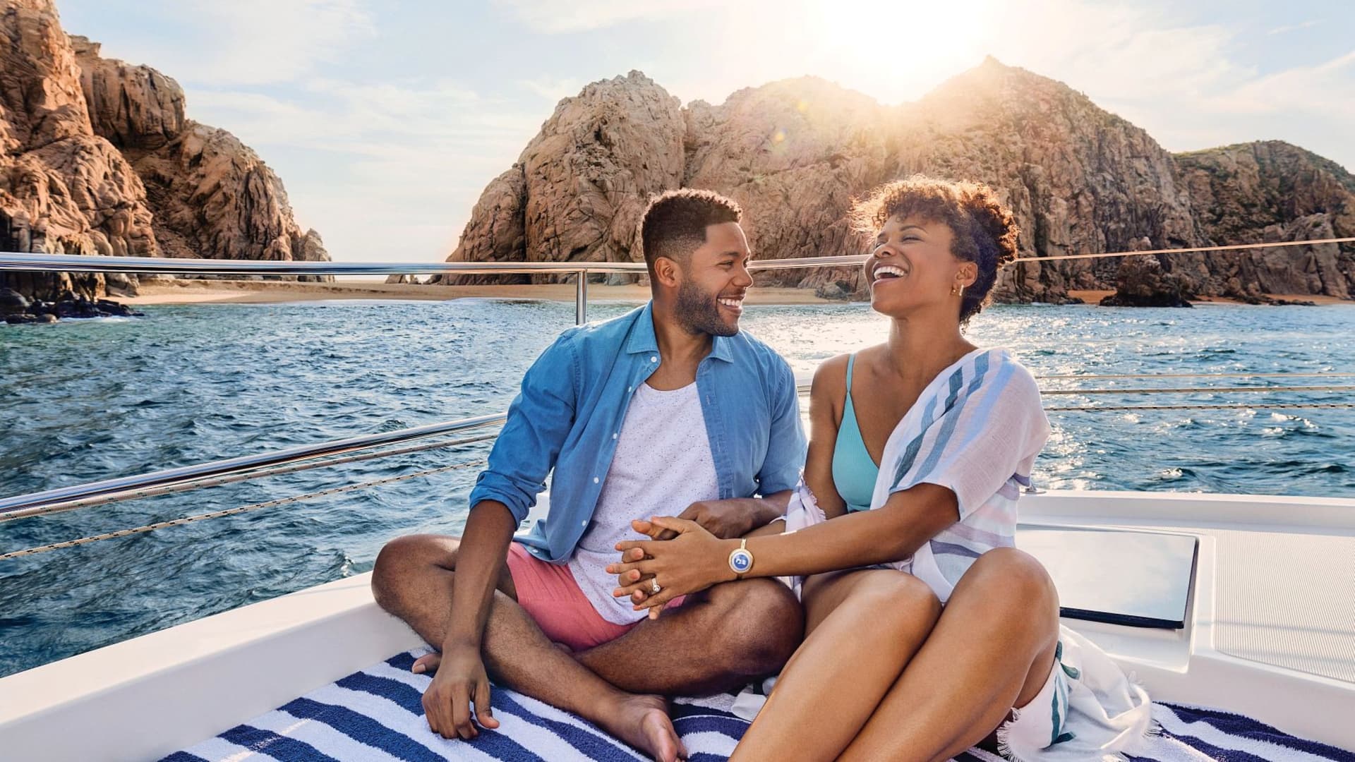 A couple laughs together on the deck of a Princess cruise ship, with sunlit ocean cliffs in the background.