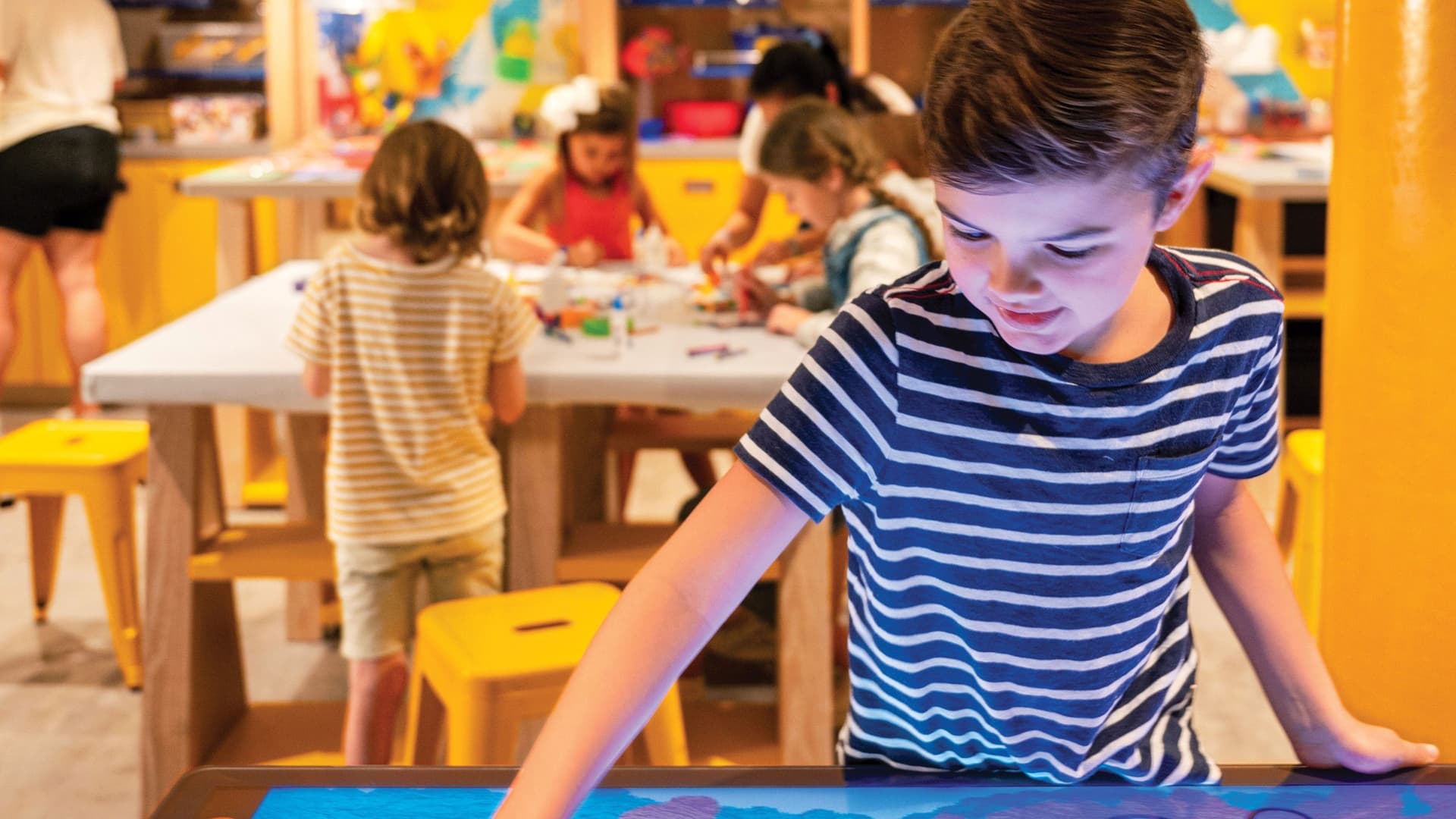 Close up of a boy playing on a table with a touchscreen, while other children engage in arts in crafts at a table in the background.