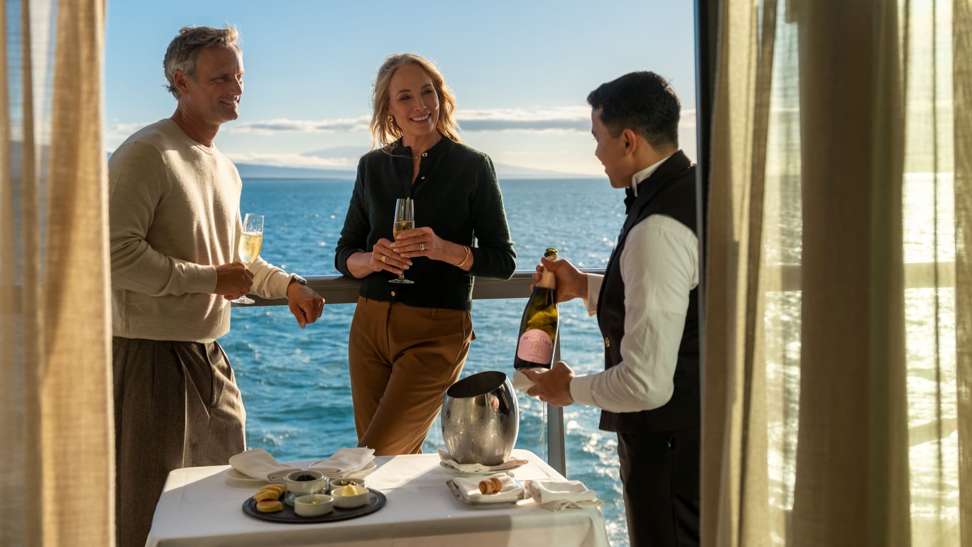 The image shows a butler in a formal uniform pouring champagne for a couple on the private veranda of a Silversea suite. The balcony overlooks a calm, blue ocean under a clear sky, with land visible in the distance. A small table is set with appetizers, a chilled ice bucket, and white linens, illustrating the personalized service and ultra-luxury atmosphere characteristic of Silversea Cruises.