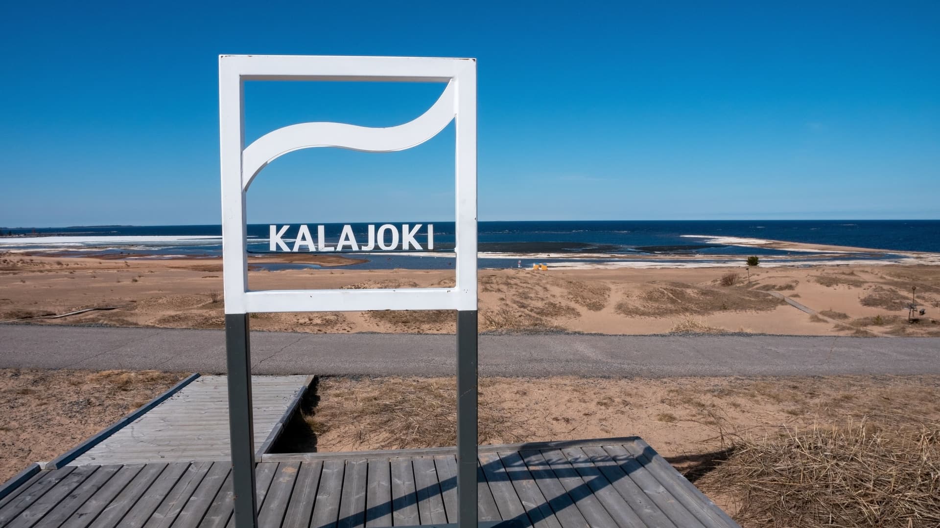 A white Kalajoki photo frame on a wooden platform overlooking a sandy beach and bright blue sea under a clear Finnish sky.