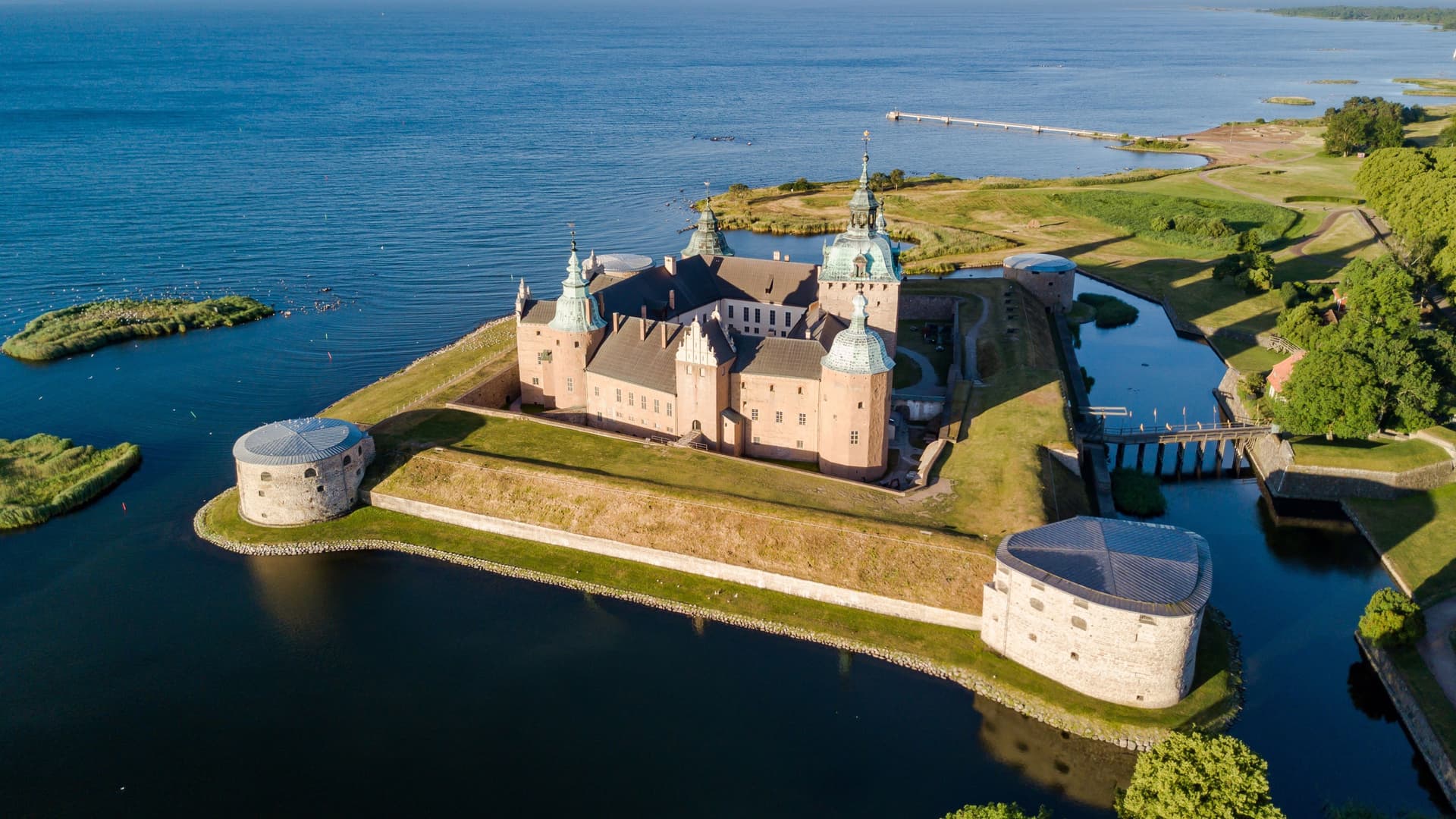 An aerial view of Kalmar Castle on its island fortress, with copper‑roofed towers, stone walls, and surrounding coastal waters stretching toward grassy shoreline and open sea.