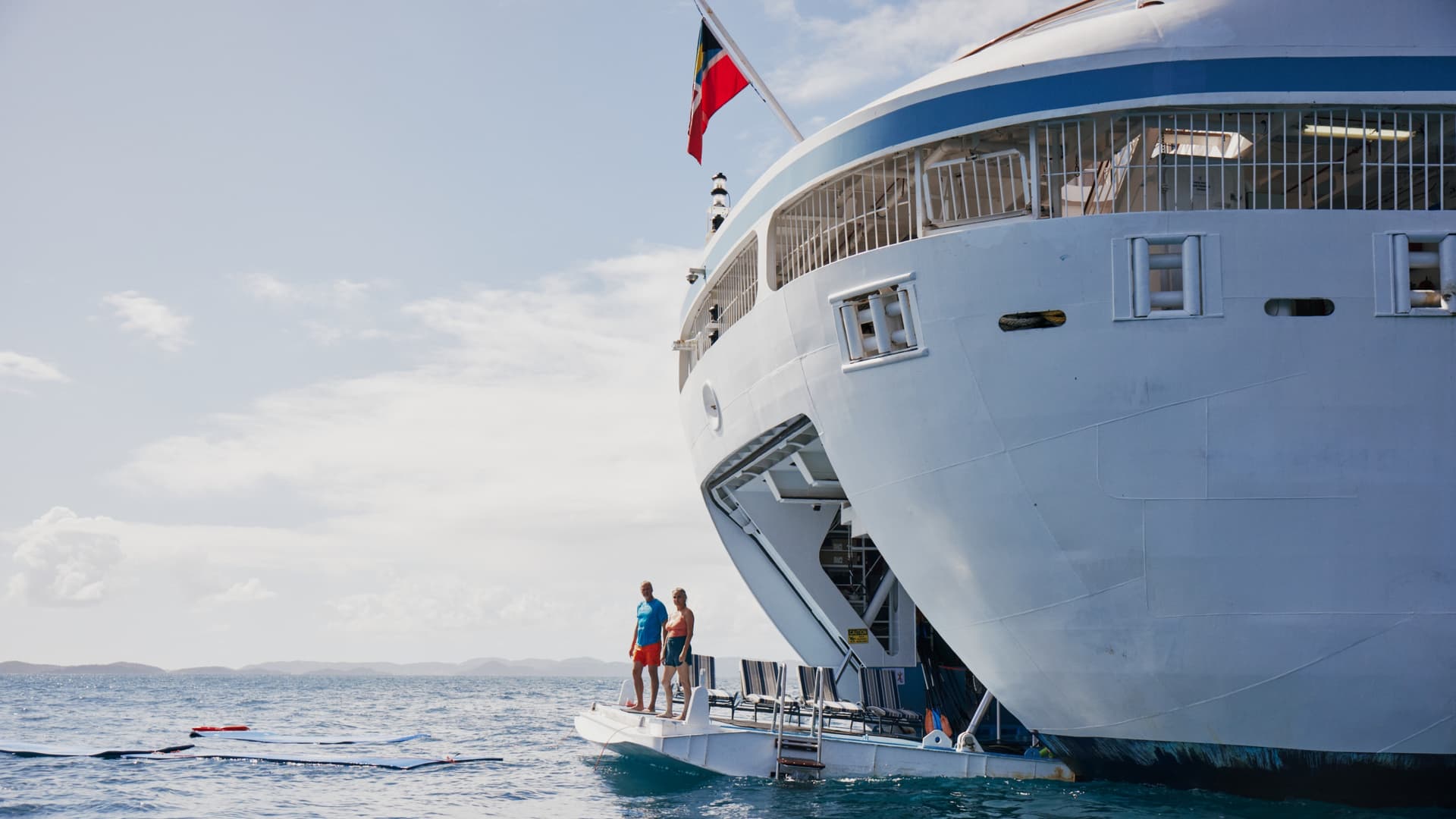 A man and woman stand on a cruise ship's retractable watersports marina platform, looking out over the open ocean where floating mats drift nearby.