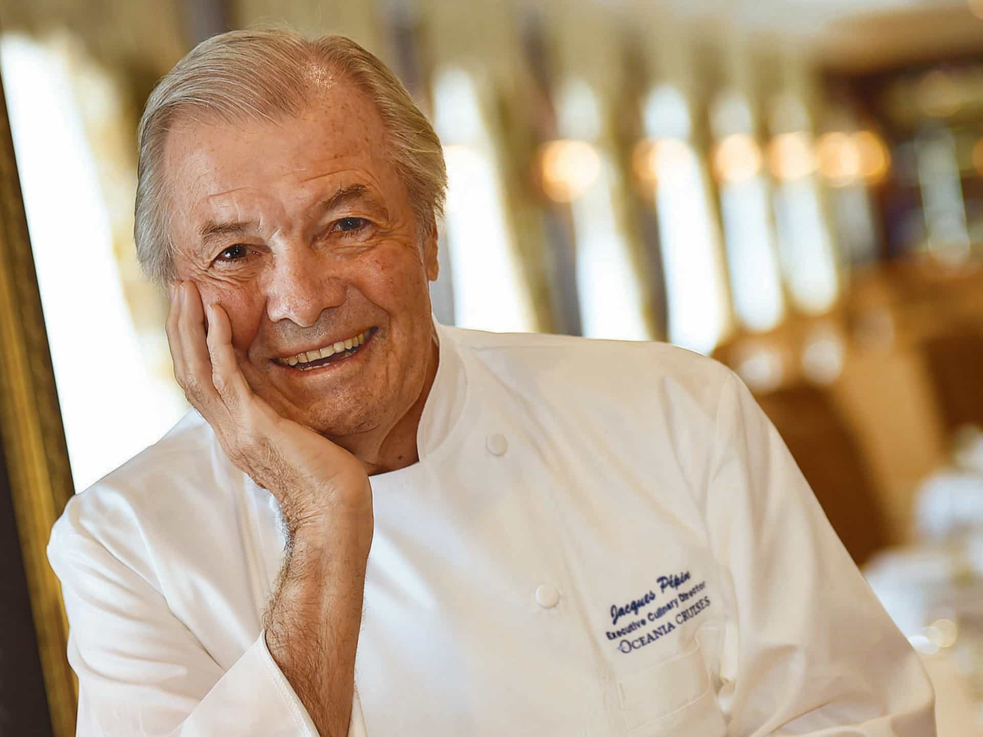 Jacques Pépin, Executive Culinary Director of Oceania Cruises, smiling in his embroidered chef whites in one of the ship's elegant dining rooms.
