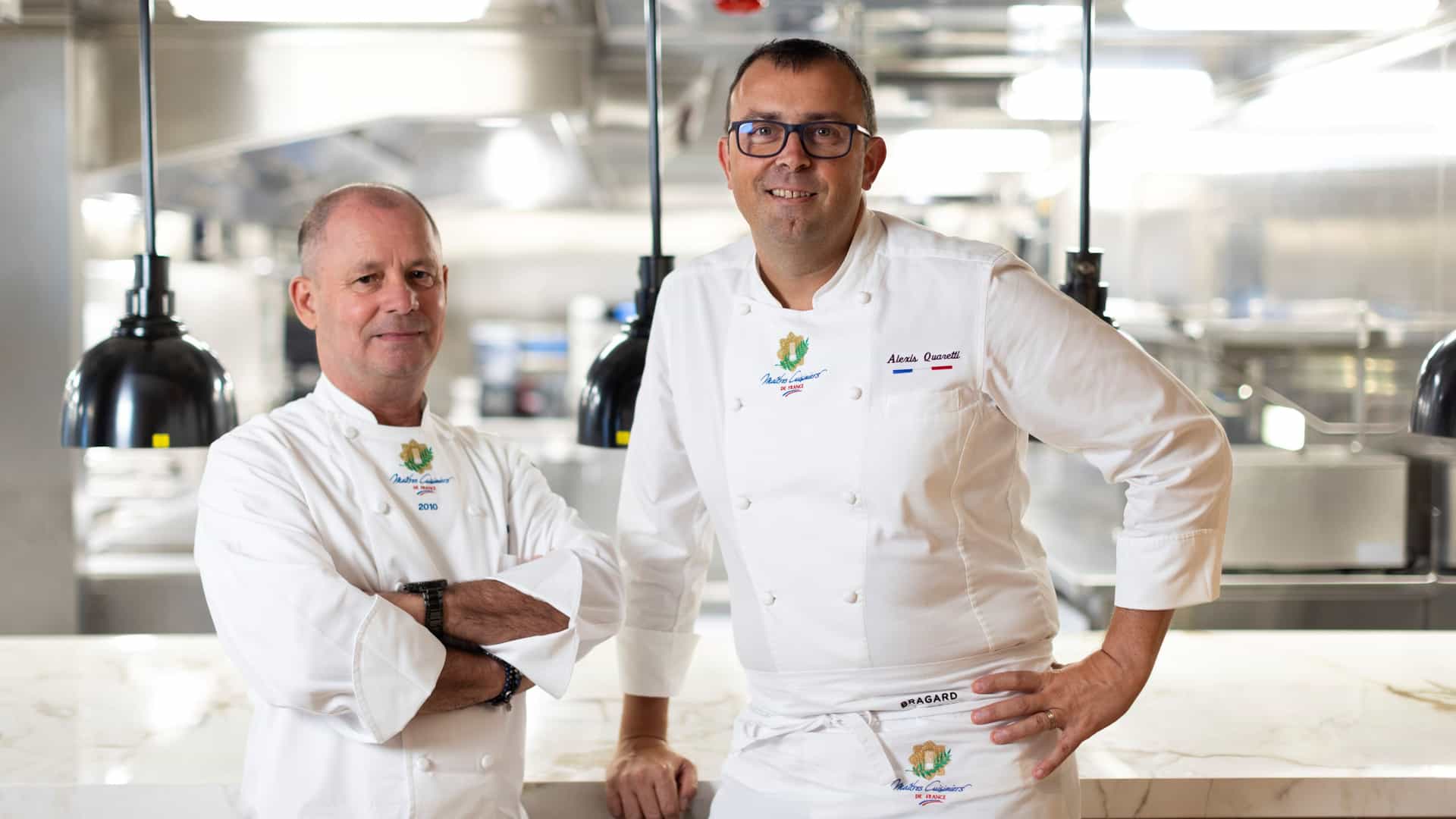 Oceania Cruises Executive Culinary Directors Eric Barale and Alexis Quaretti in chef whites bearing the Maîtres Cuisiniers de France crest, posing in a ship's professional galley kitchen.